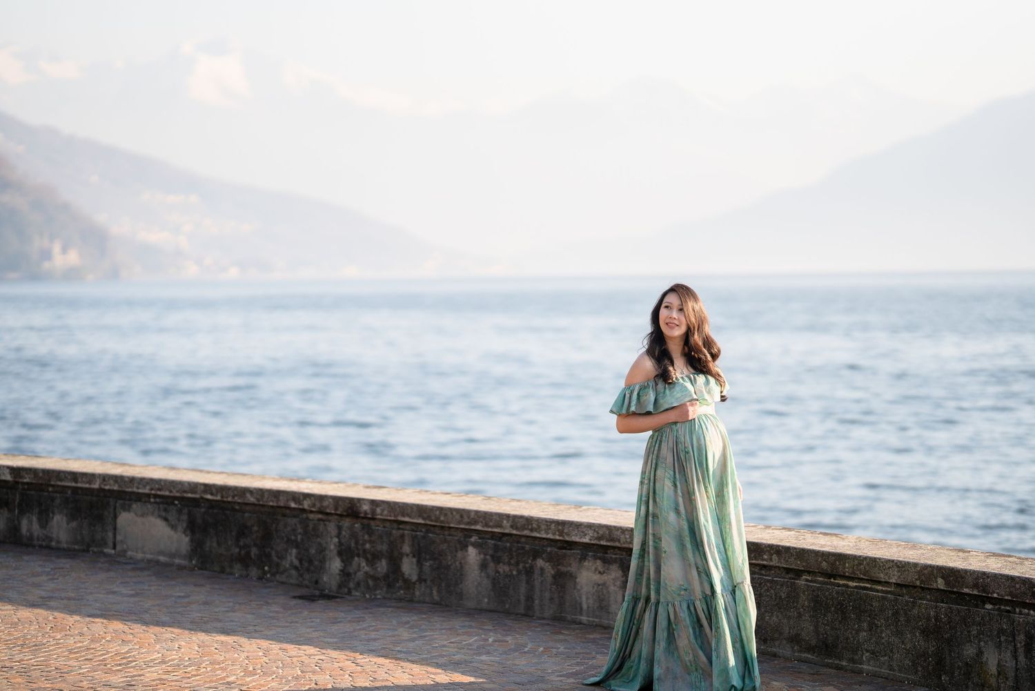 Woman in elegant teal gown poses on lakeside promenade with misty mountains and serene water in background.