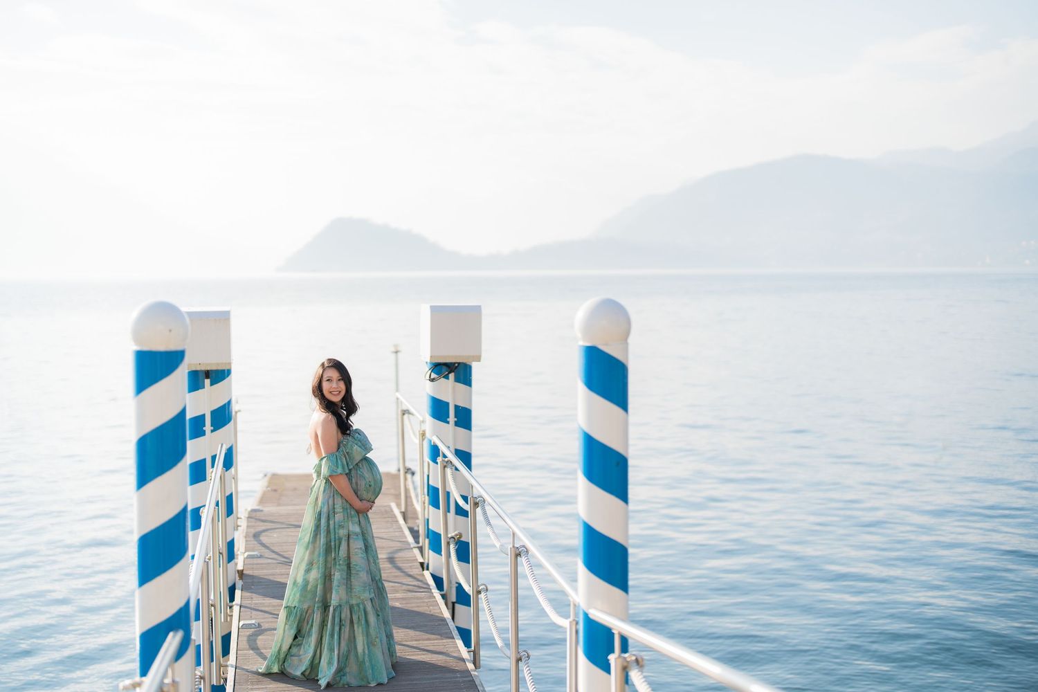 A woman in a flowing green gown stands on a blue-white striped pier overlooking a calm blue lake with mountains.