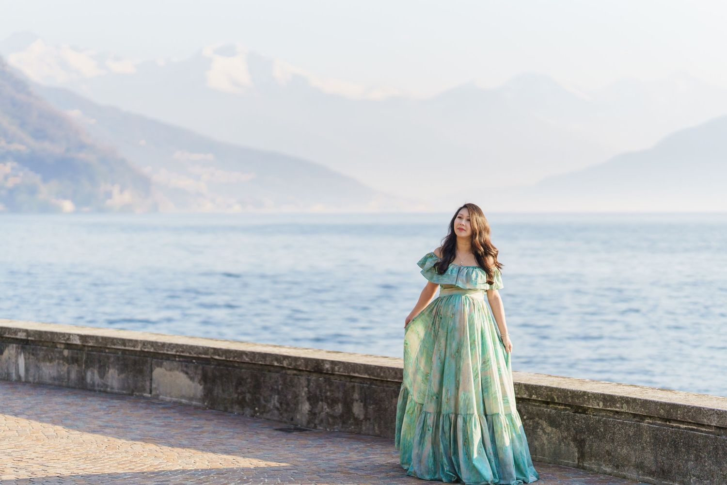 Woman in teal off-shoulder gown poses by lakeside promenade with misty mountains in background.