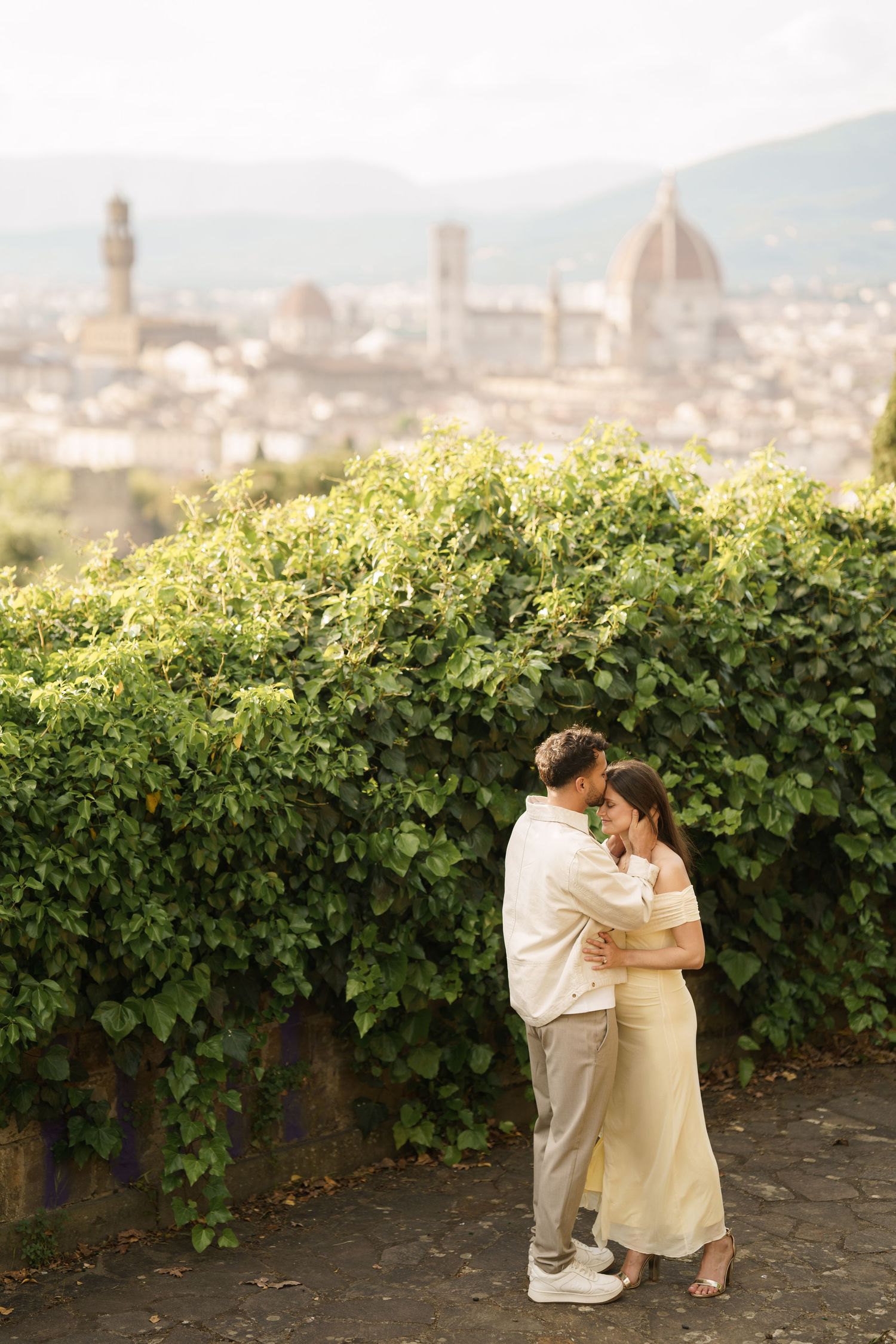 Romantic couple embracing in front of lush ivy wall with Florence skyline and Duomo cathedral in background.