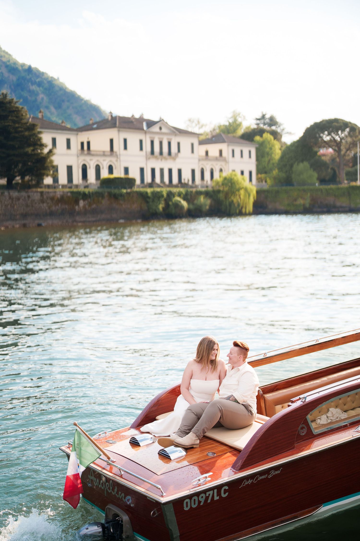 Couple sitting on a classic wooden boat on Lake Como, Italy, with a historic villa and mountains in the background.