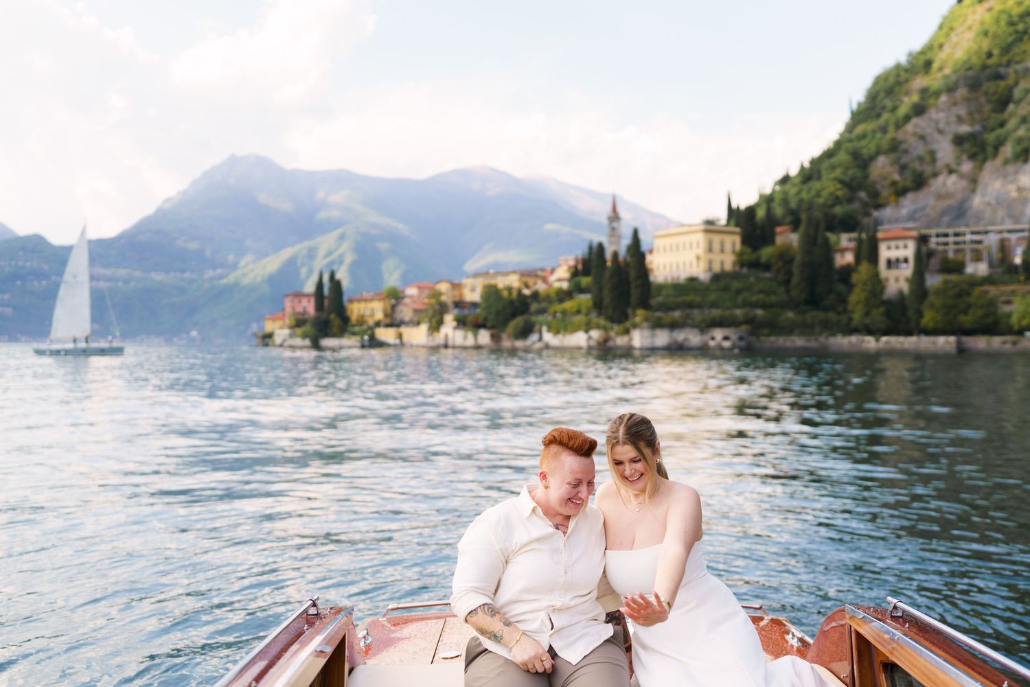 Couple in wedding attire sitting together on a wooden boat on Lake Como, Italy, with mountains and village in background.
