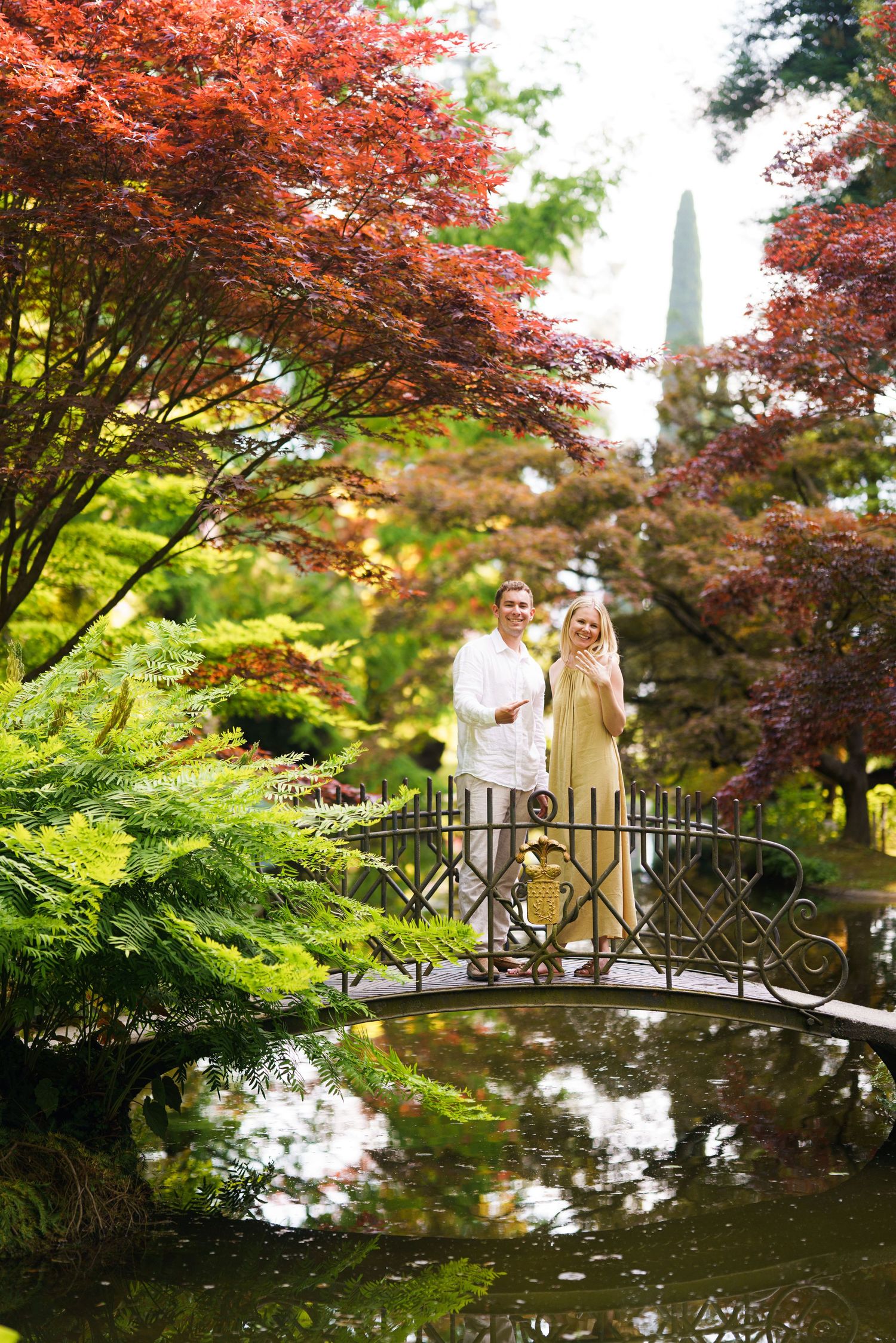 Couple standing on ornate iron bridge over pond in lush Japanese garden with red maple trees.