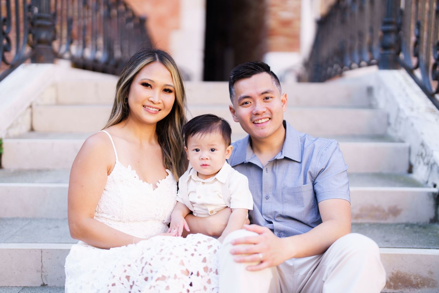 Happy Asian family with parents and baby sitting on outdoor steps, smiling in a bright casual photo session.