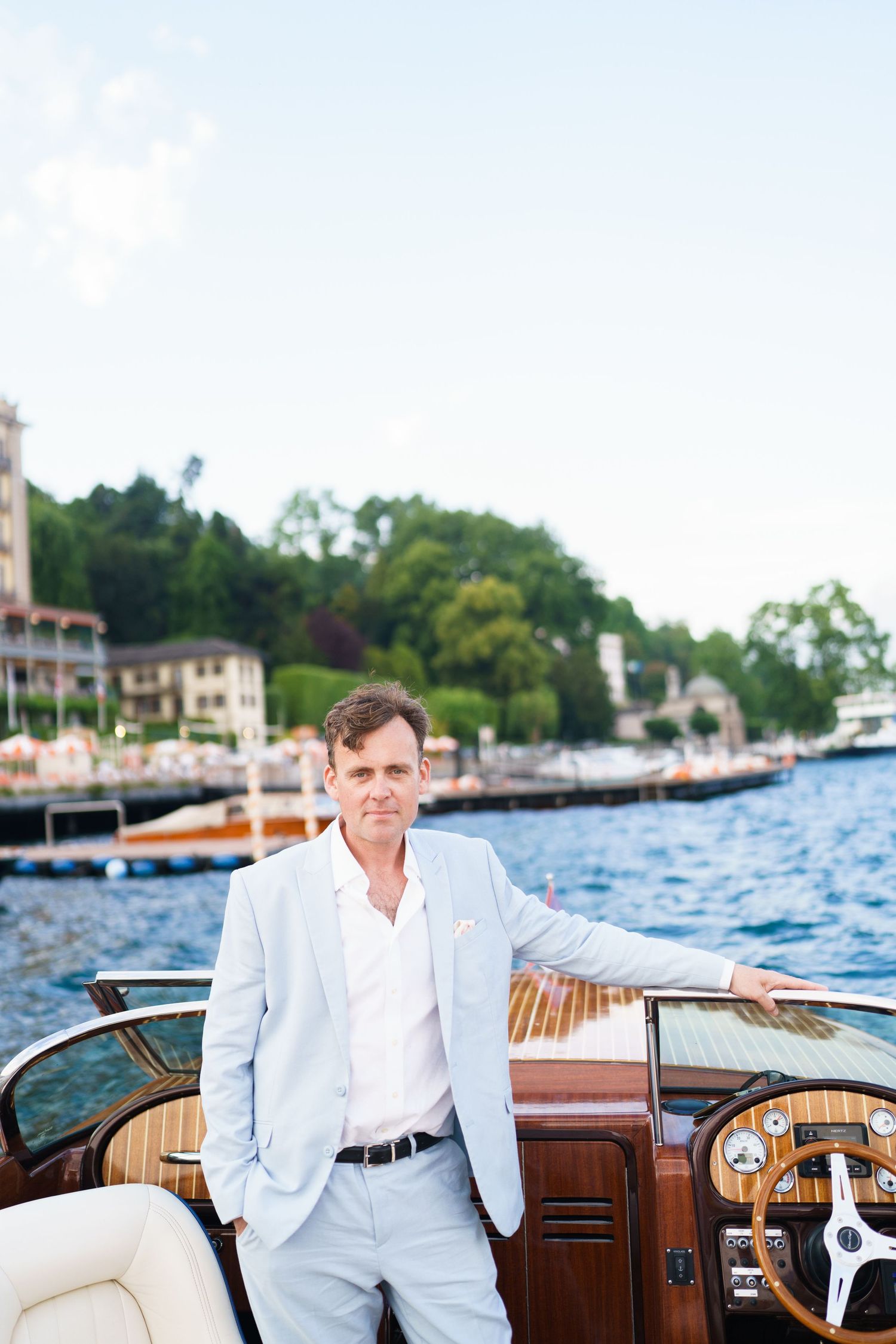 Elegant man in light blue suit posing on a classic wooden boat on a serene lake with waterfront buildings.