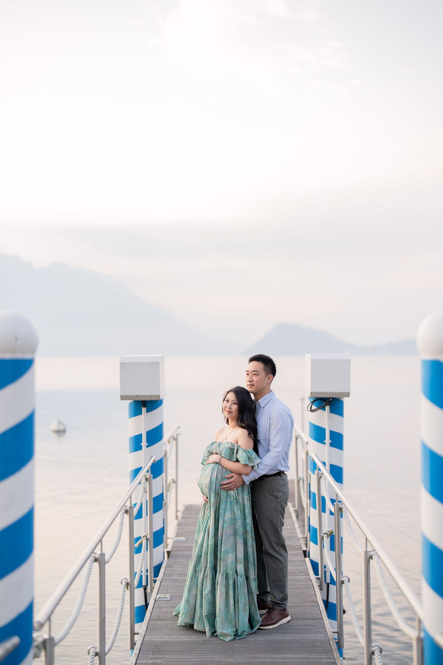 Pregnant couple embracing on a pier overlooking calm water and mountains, woman in teal floral gown, man in blue shirt.