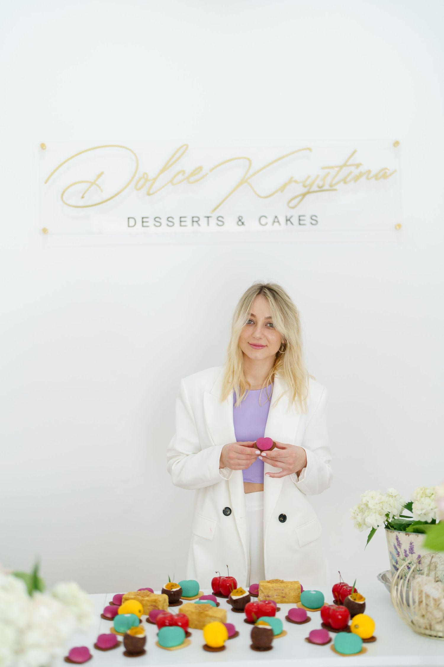 Blonde woman in white blazer holding macaron at Dolce Krystina Desserts & Cakes table with colorful macarons and pastries.