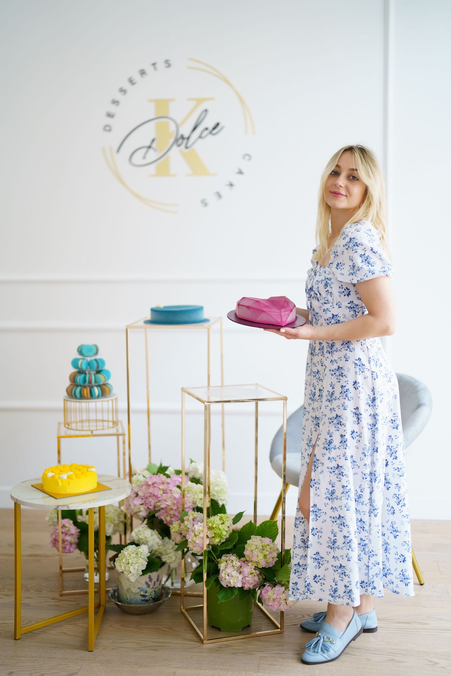 Blonde woman in floral dress holding pink cake at Dolce K Desserts shop with flowers and colorful cakes on gold stands.