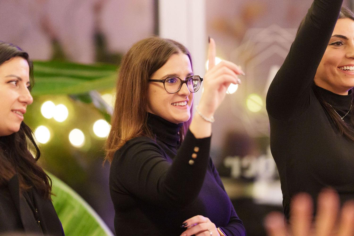 Three women at an event, one smiling and pointing upward, wearing glasses and a black turtleneck.