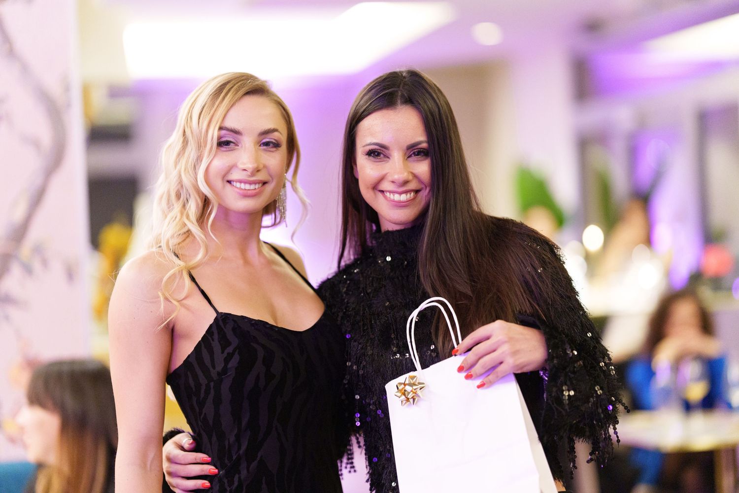 Two elegantly dressed women smiling together at an event, one blonde in black dress, one brunette holding a white gift bag.