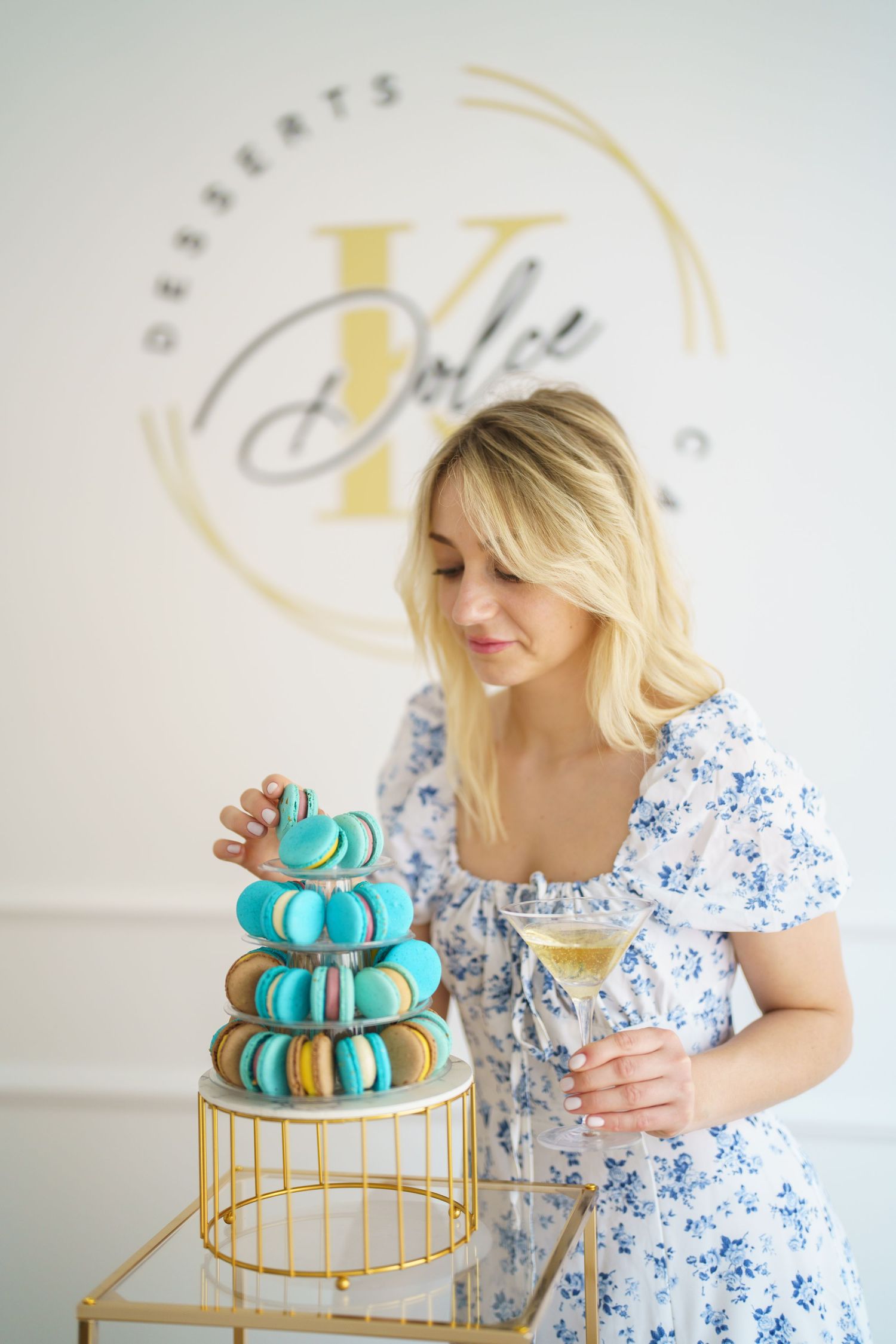 Woman in floral dress arranging colorful macarons tower display at Dolce Desserts cafe holding champagne glass.