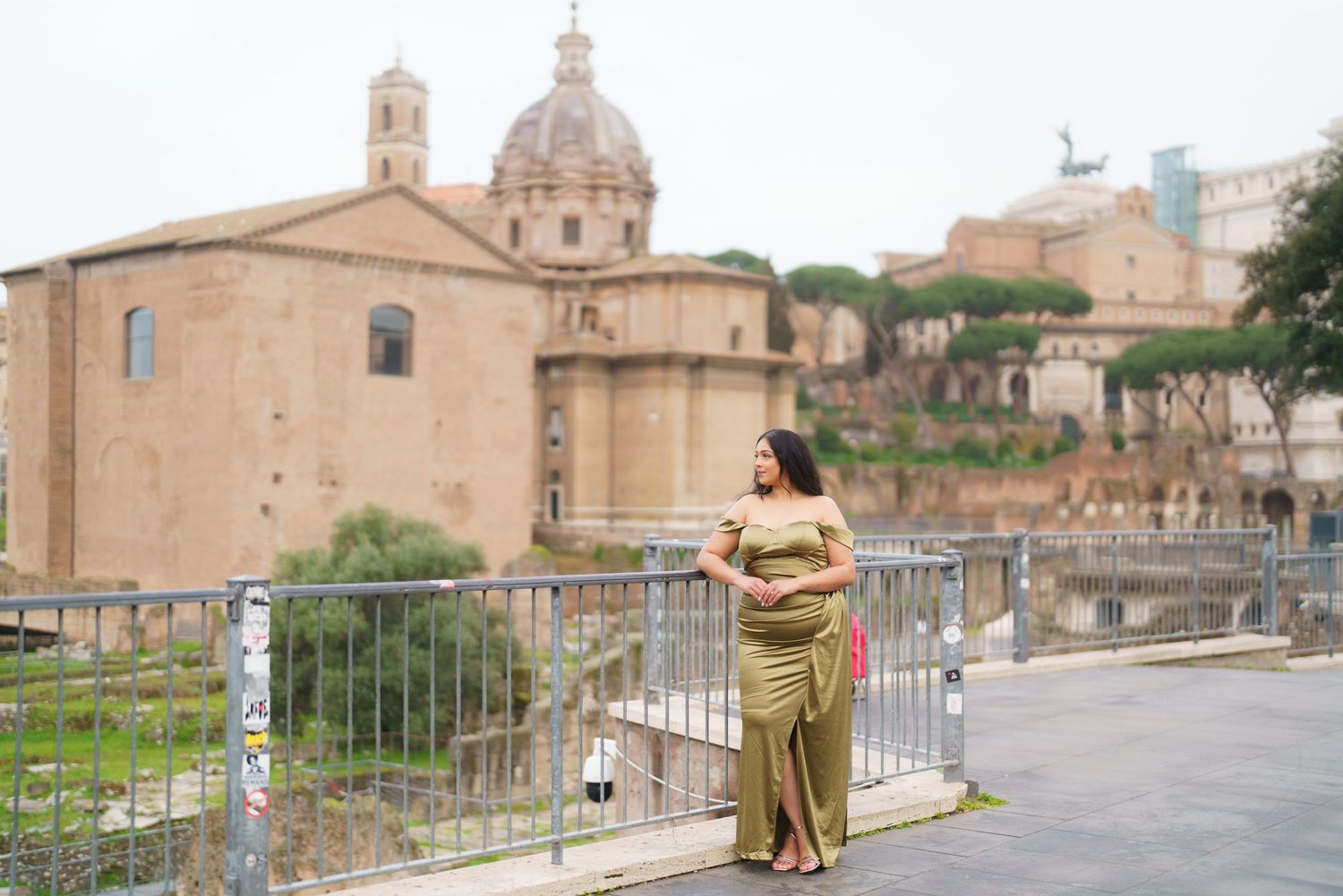 Pregnant woman in gold dress poses by Roman Forum ruins with ancient church dome backdrop in Rome, Italy.
