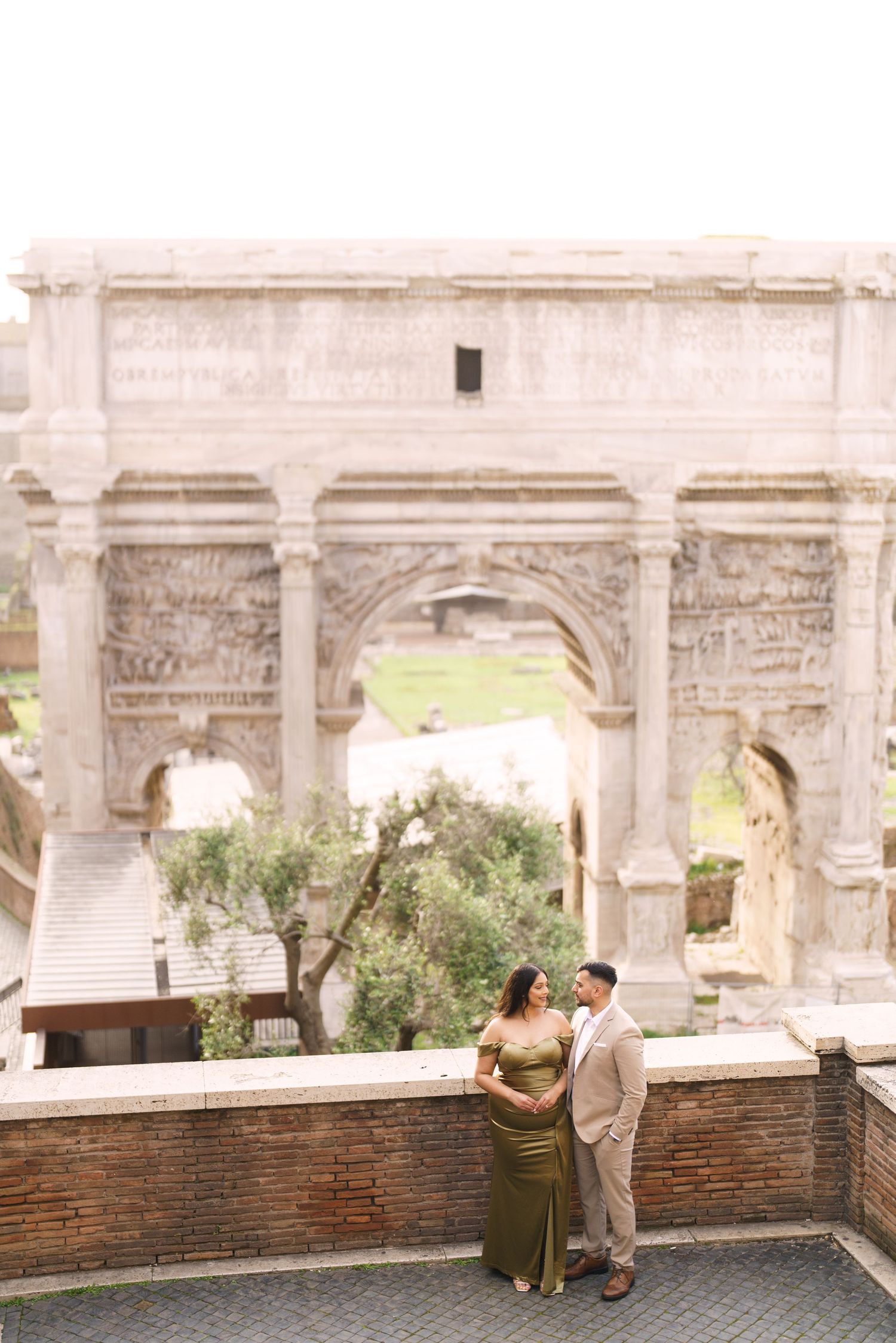 Couple poses in elegant attire before the Arch of Septimius Severus in Rome, Italy.