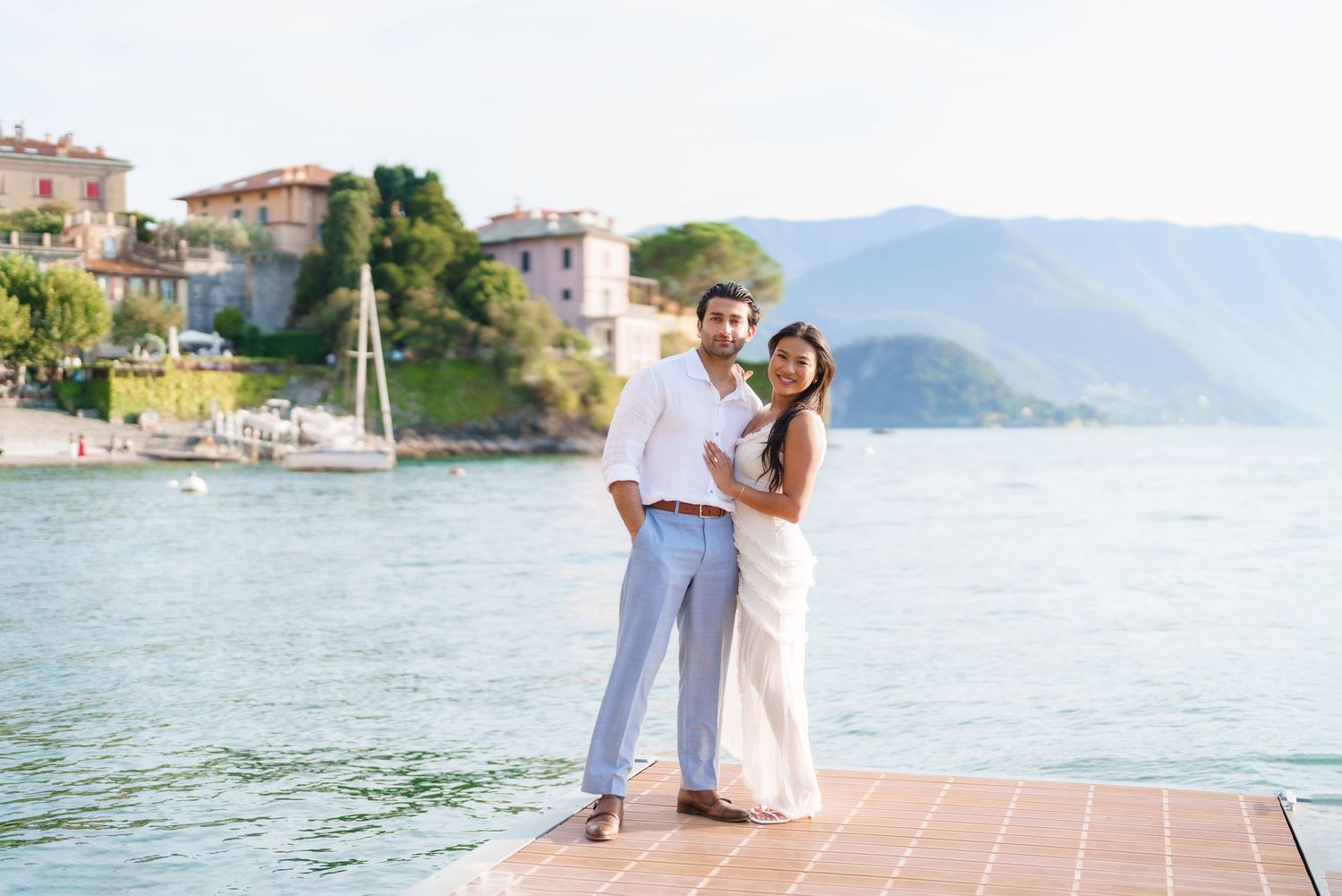 Couple in white outfits posing on a dock with Lake Como and mountain backdrop in Italy.