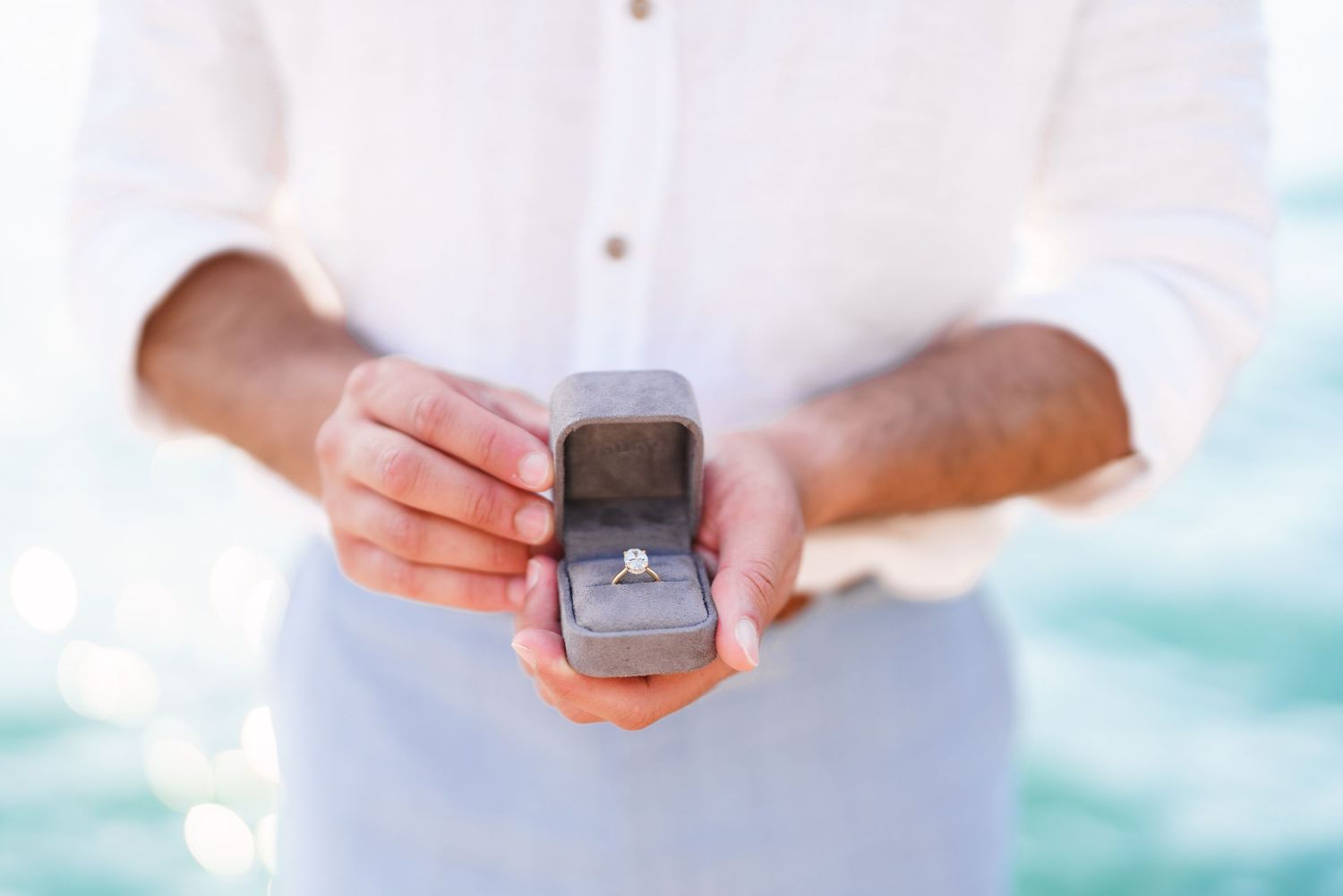 Man in white shirt holding open ring box with diamond engagement ring outdoors near ocean.
