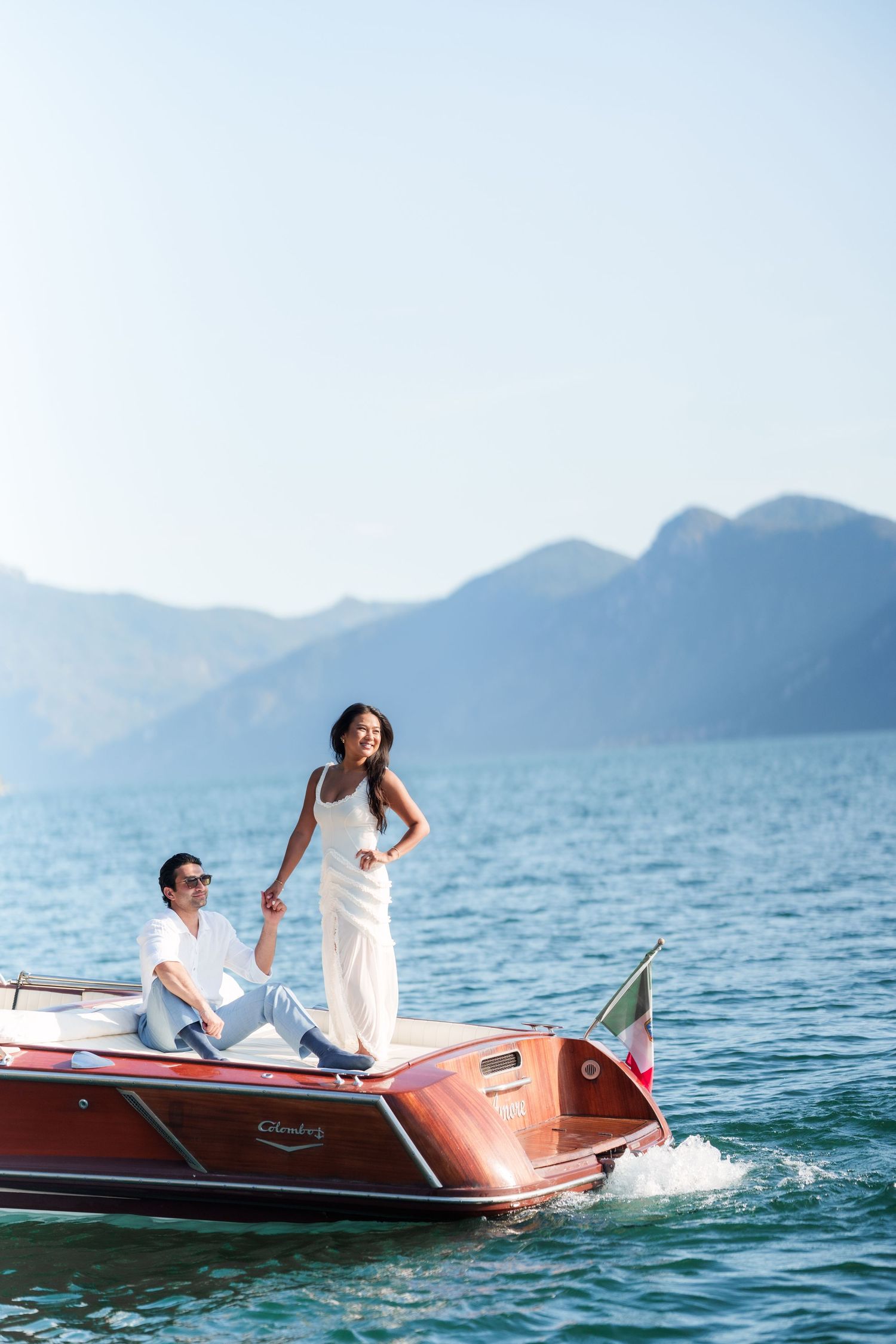Couple in white attire on a classic wooden speedboat with Italian flag, mountains and blue water in background.