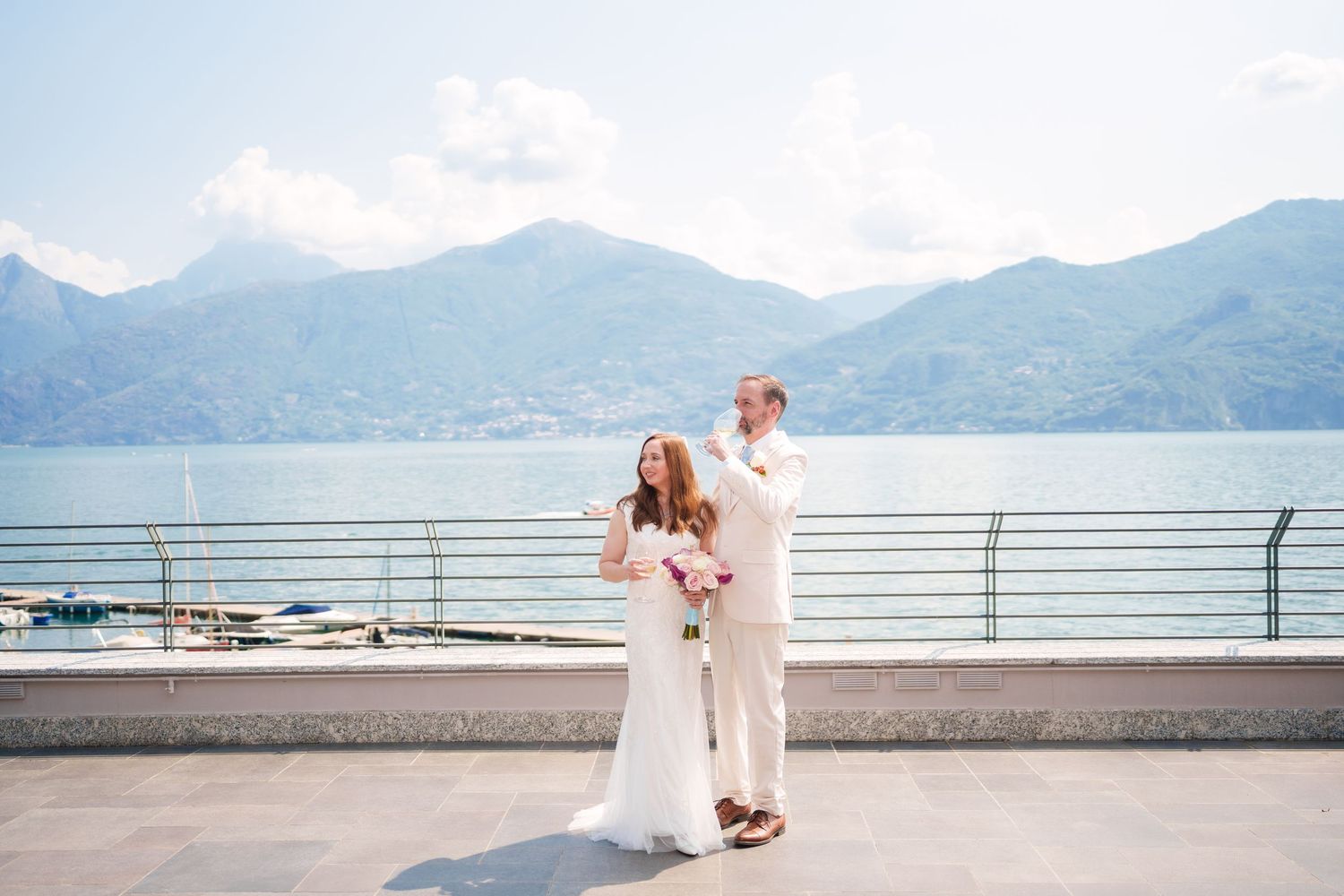 Bride and groom pose on lakeside terrace with mountain backdrop on their wedding day in Italy.