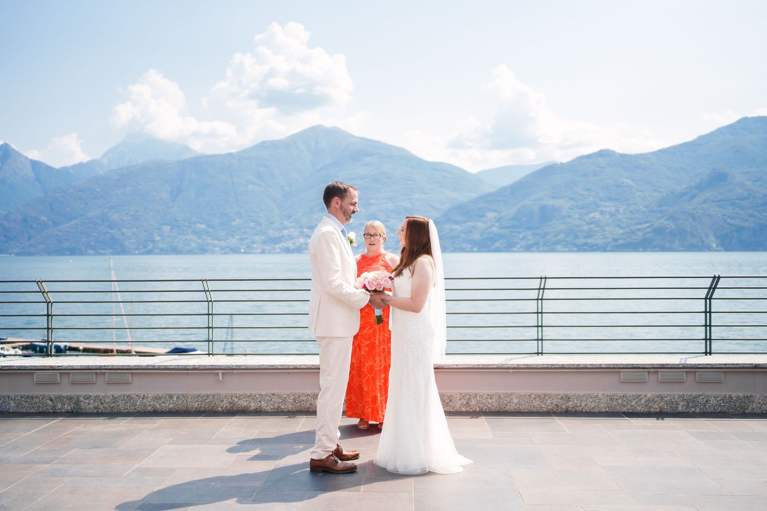 Couple exchanges vows on a scenic lakeside terrace with mountain backdrop, officiated by a woman in an orange dress.