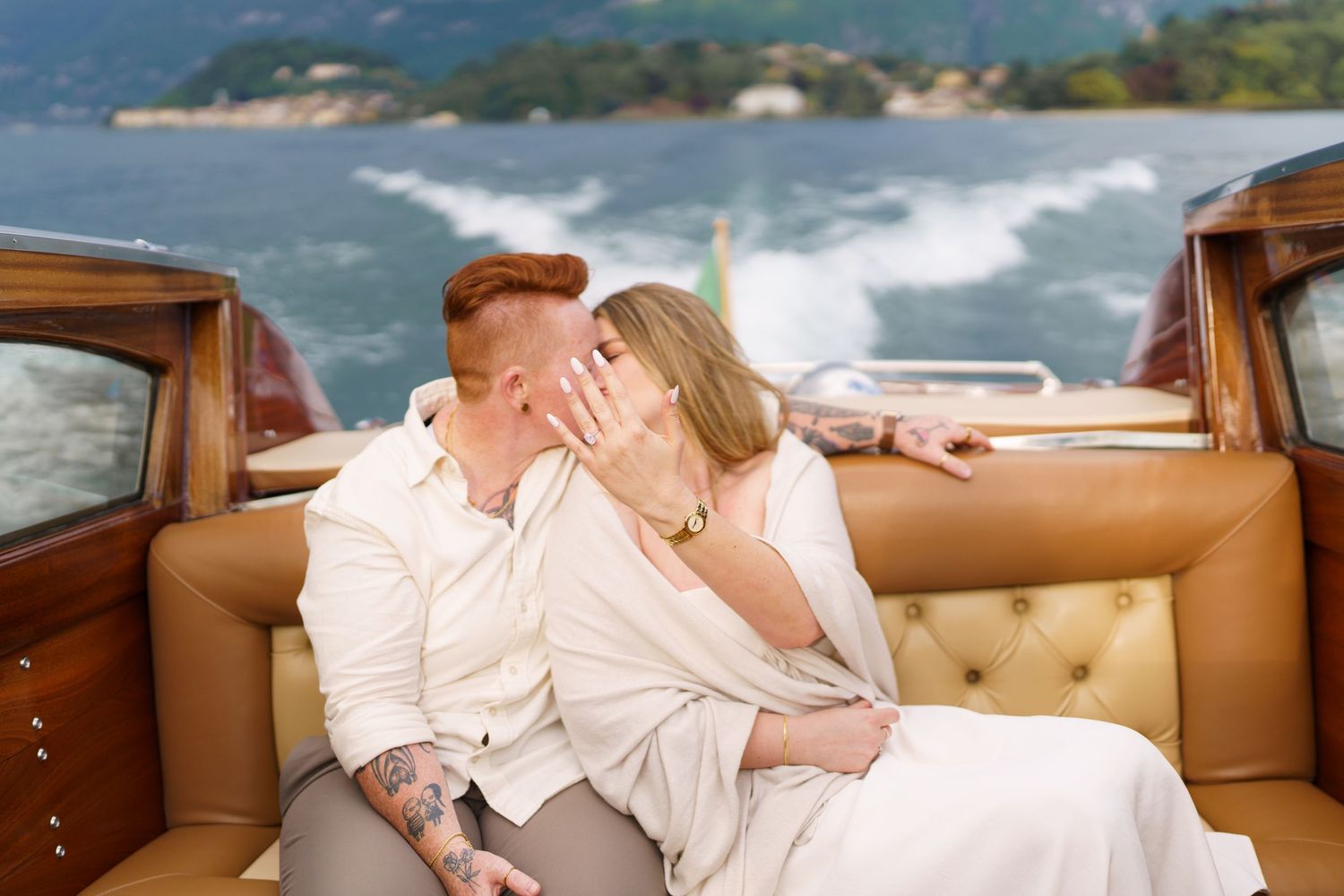 Newlyweds share a romantic kiss on a vintage wooden boat with scenic lake and mountains behind them.