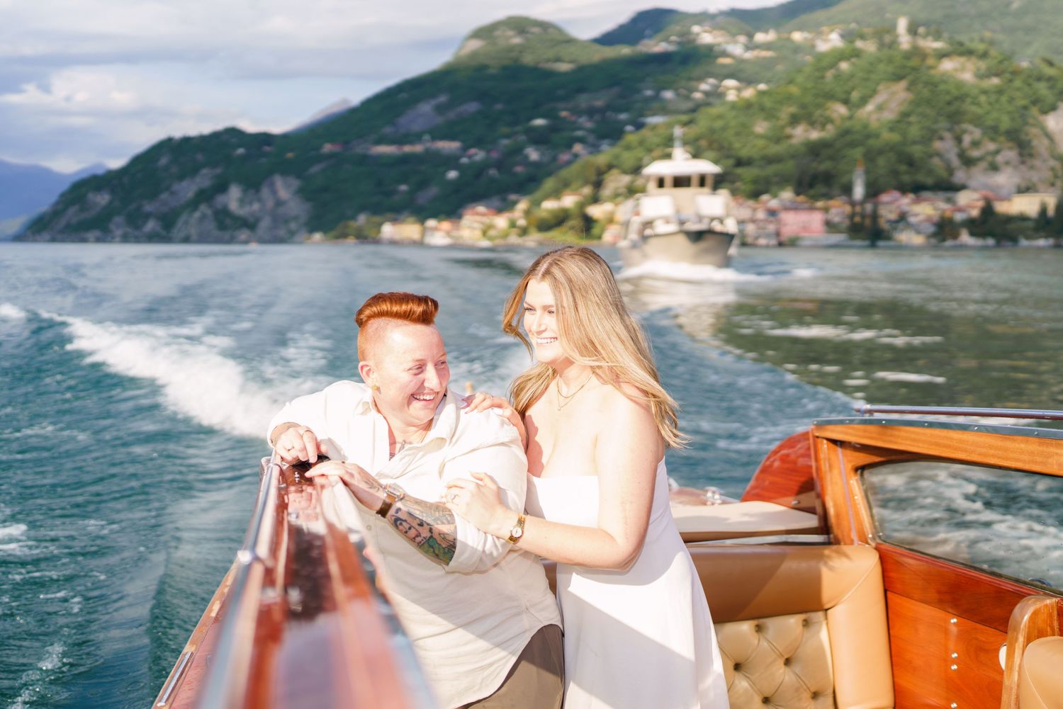 Couple laughing on a wooden boat on Lake Como, Italy, with green mountains and blue water in the background.