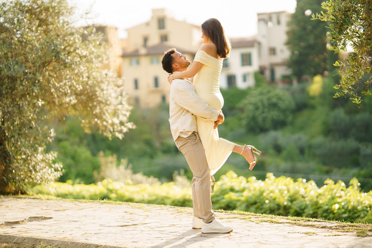 Romantic couple embracing outdoors, man lifting woman in sunny garden with lush greenery and historic buildings.