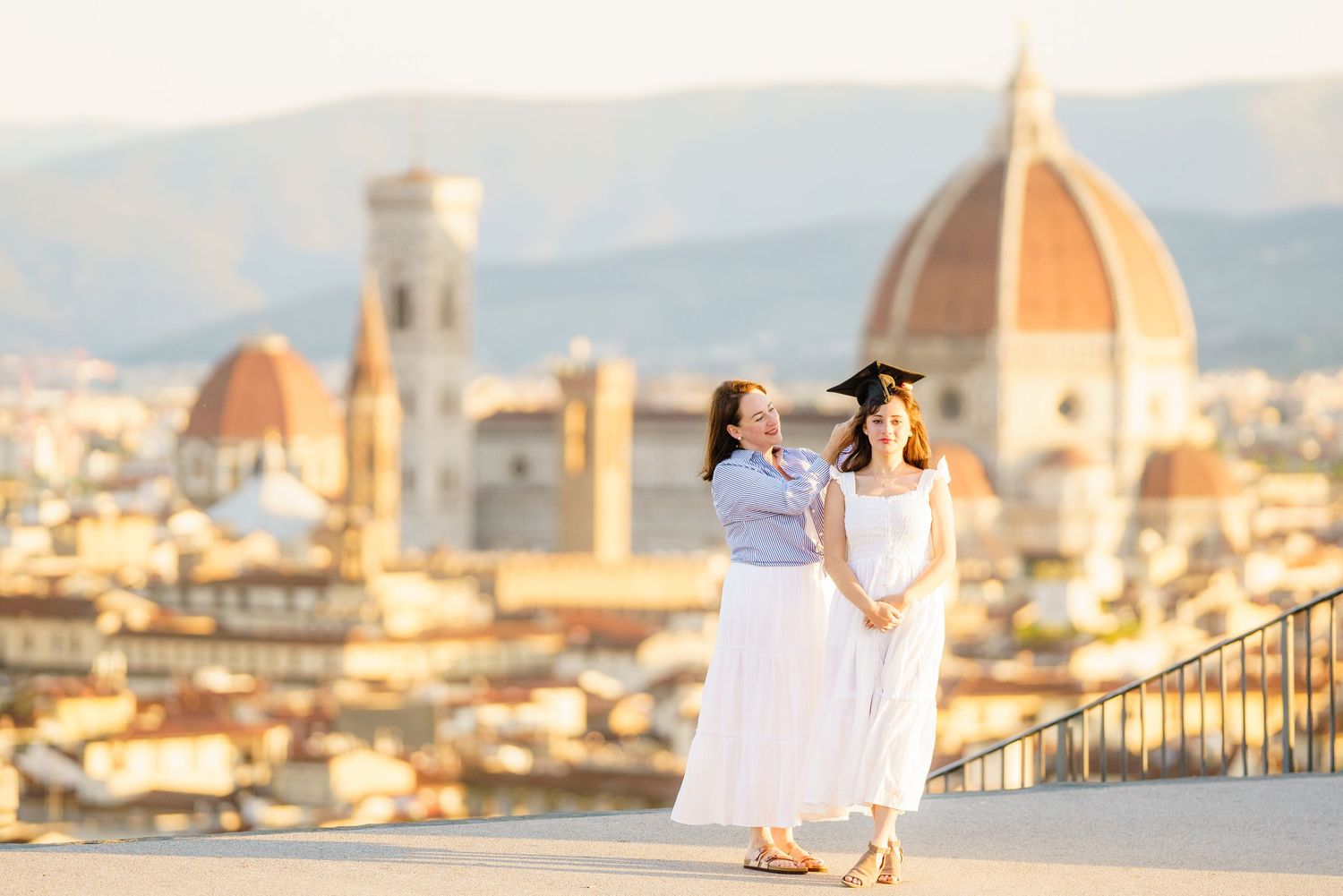 Two women in white dresses pose with Florence's iconic Duomo cathedral skyline at golden hour.