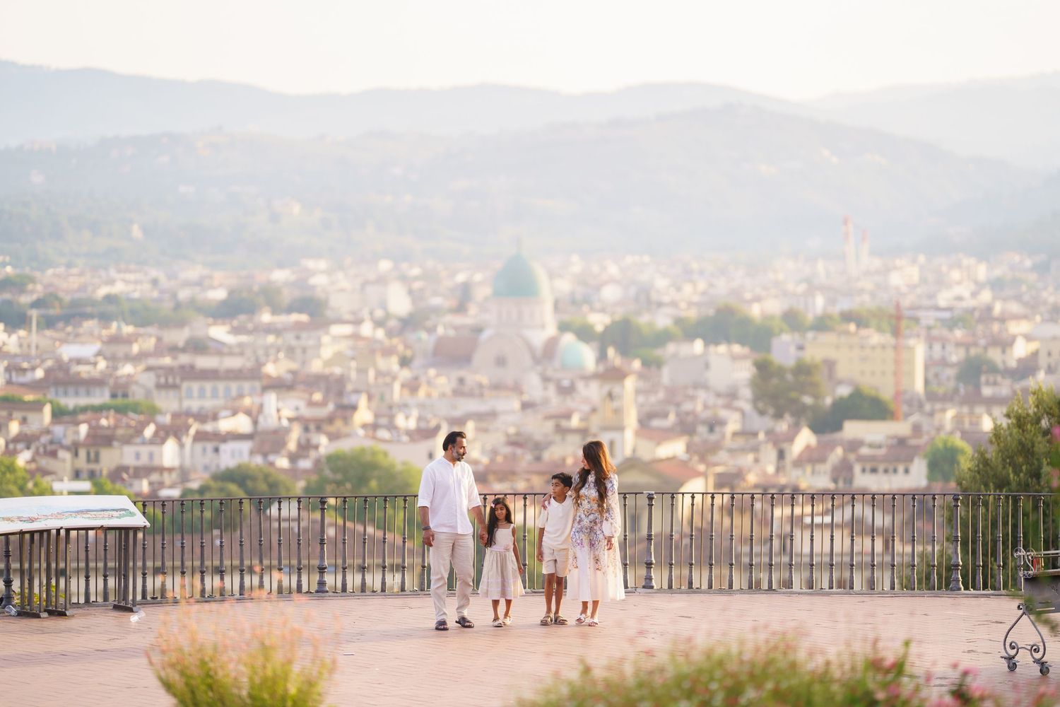 Family of four standing on a scenic overlook terrace with a panoramic cityscape and misty hills in the background.