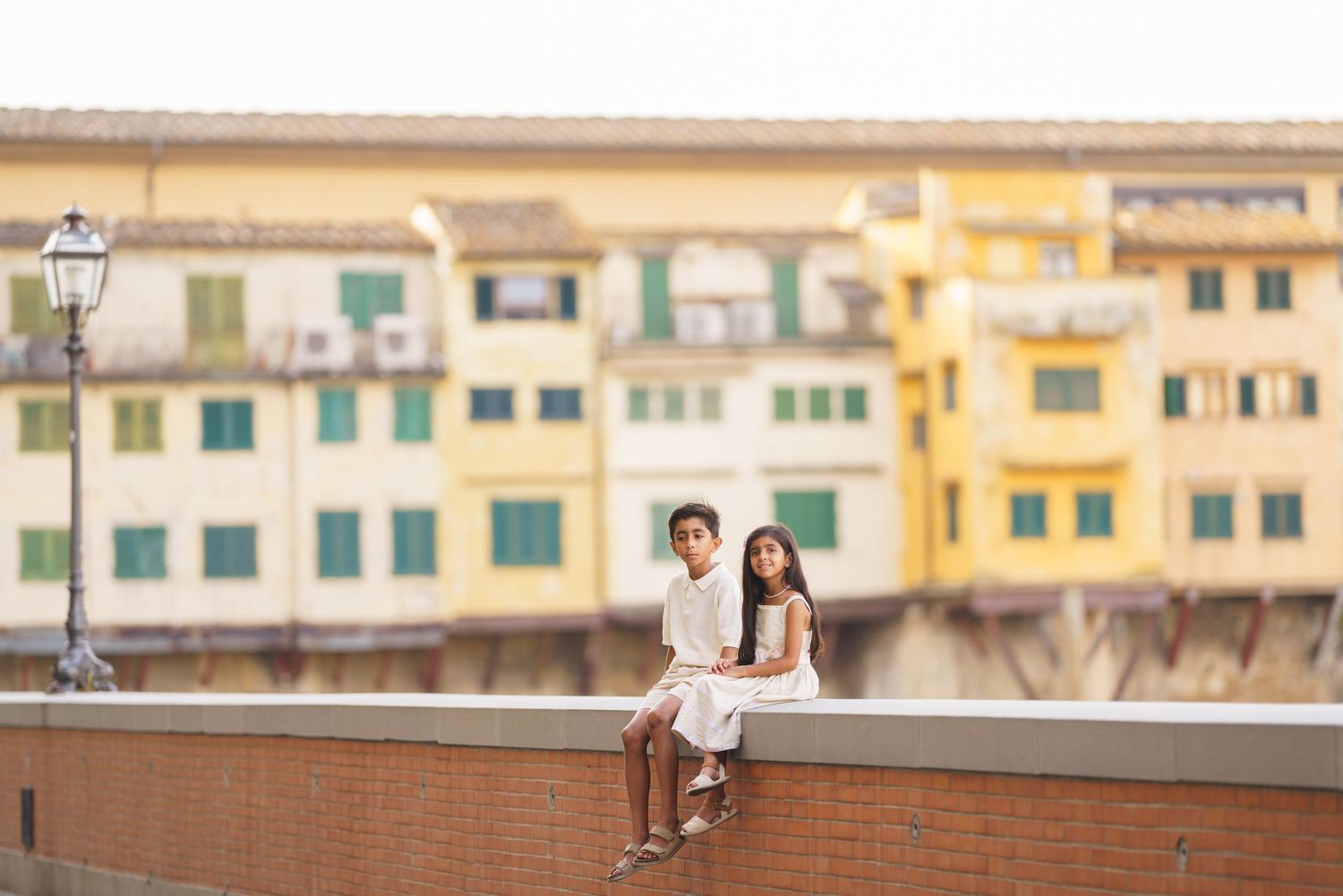 Couple in white sitting on a wall by Ponte Vecchio, Florence, with colorful historic buildings behind.