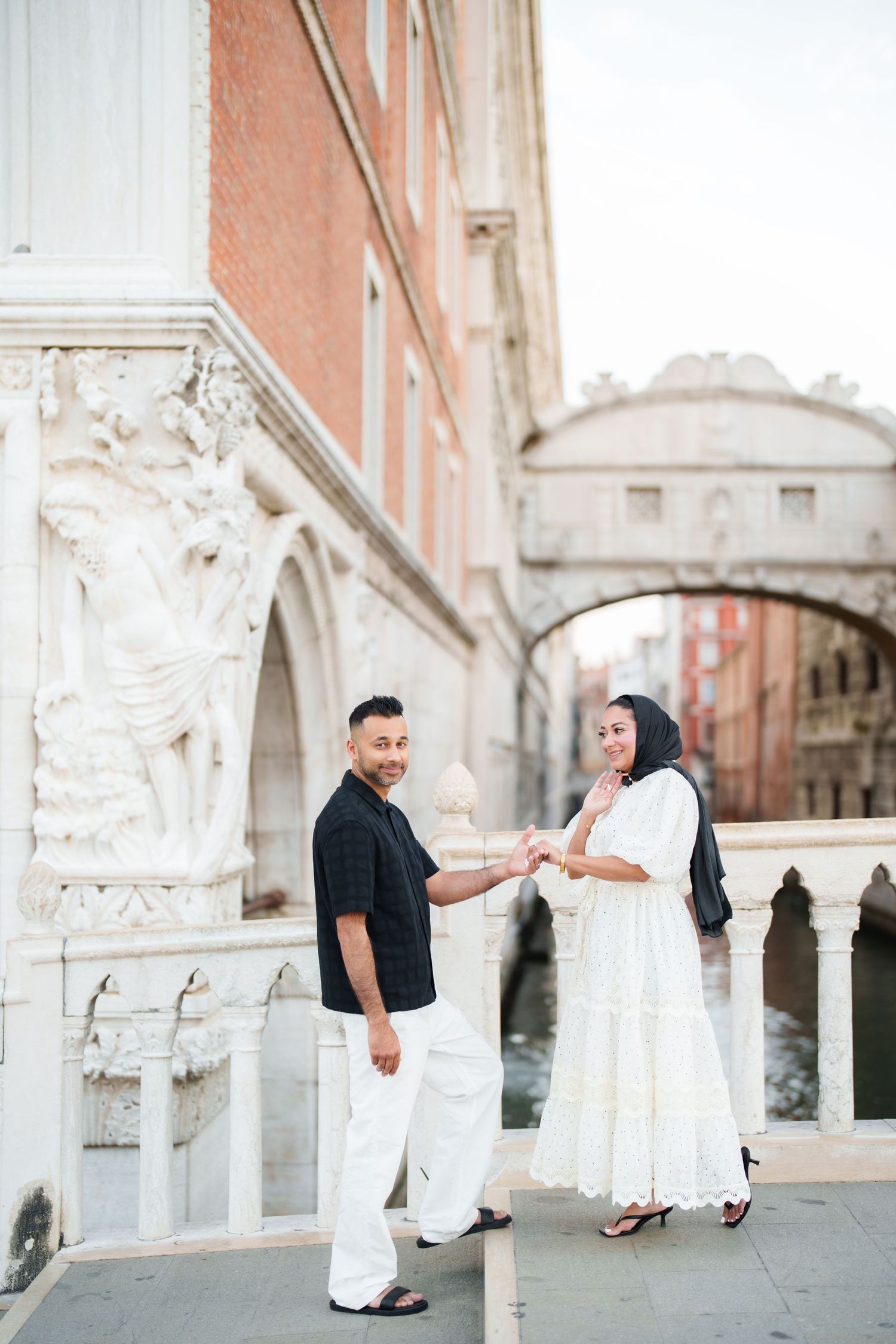Couple holding hands near the Bridge of Sighs in Venice, man in black shirt and white pants, woman in white dress.