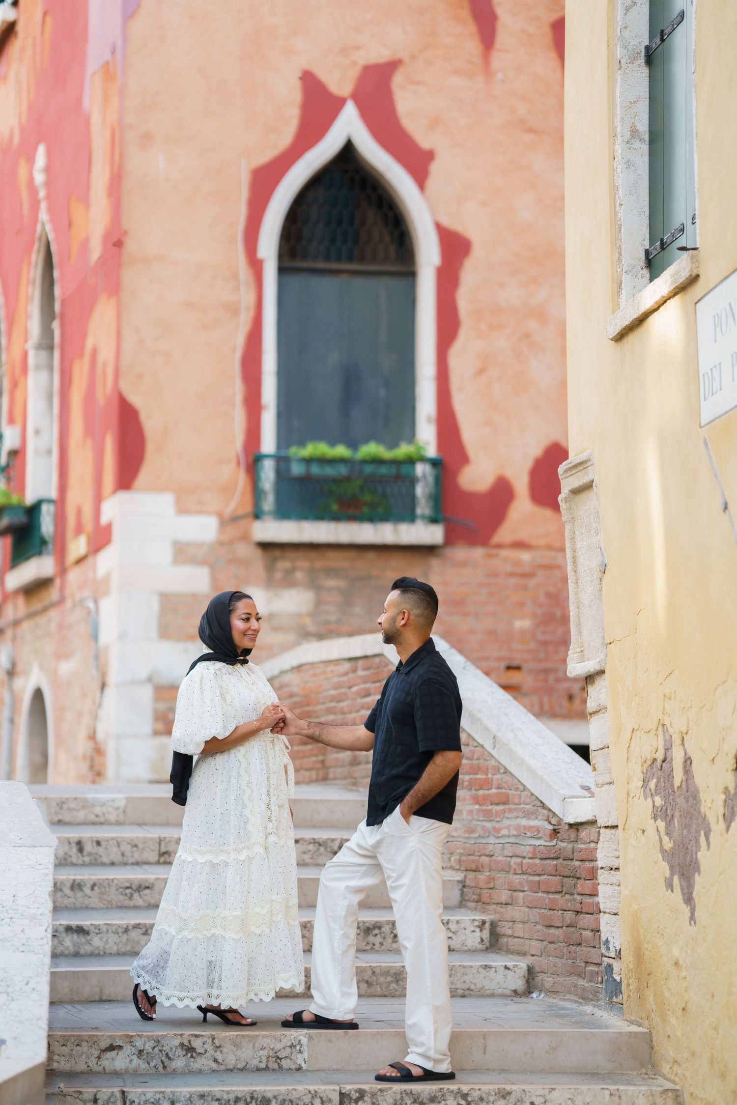 Couple in elegant attire holding hands on historic Venice steps with colorful architecture backdrop.