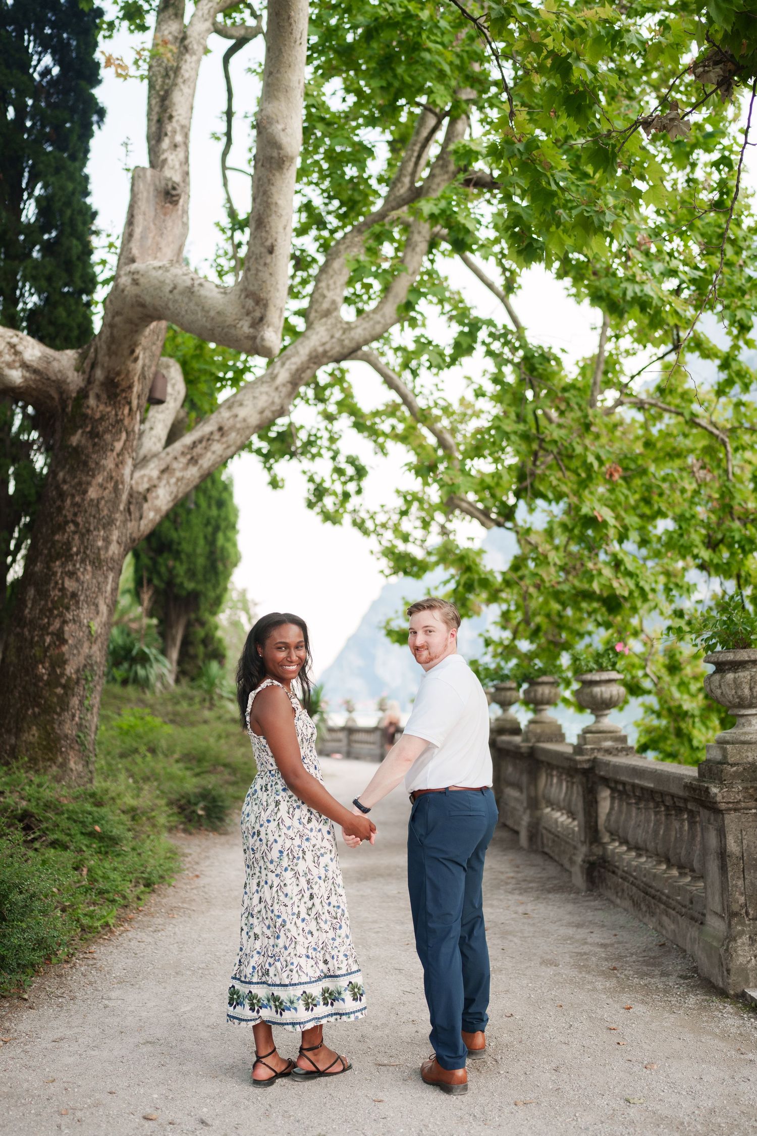 Happy interracial couple holding hands under lush green trees on a scenic Italian lakeside garden path.