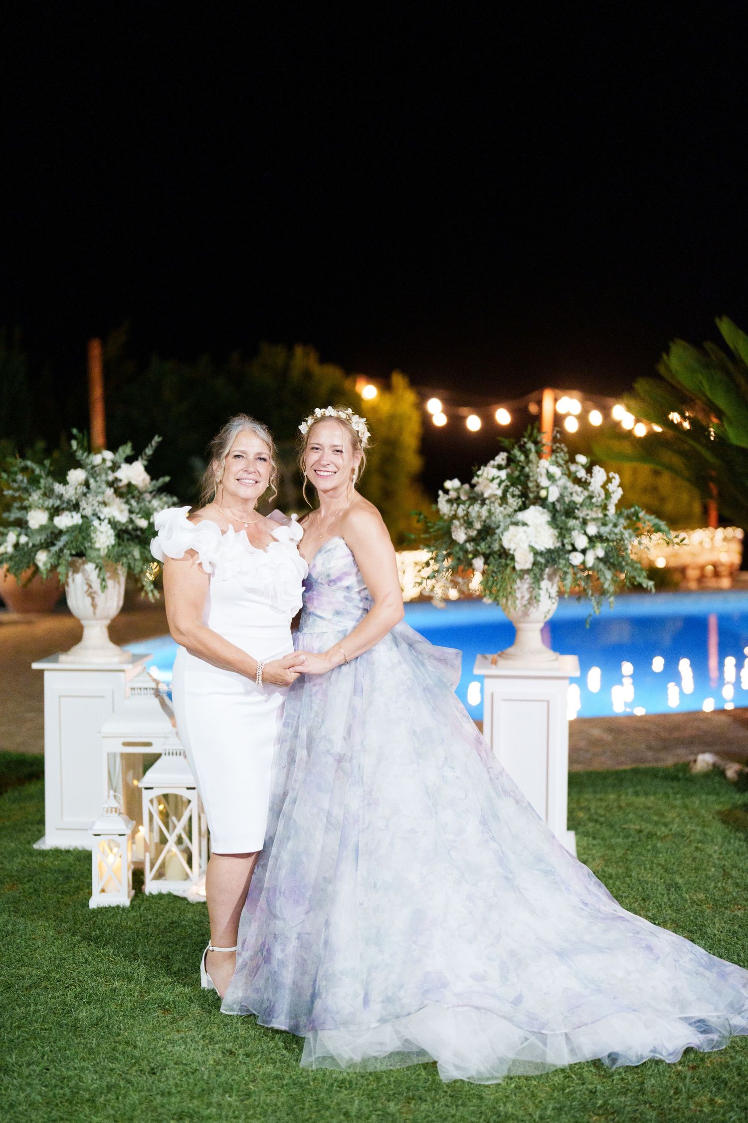 Two women pose outdoors at night near a pool, one in a white dress and one in a lavender wedding gown with floral decor.