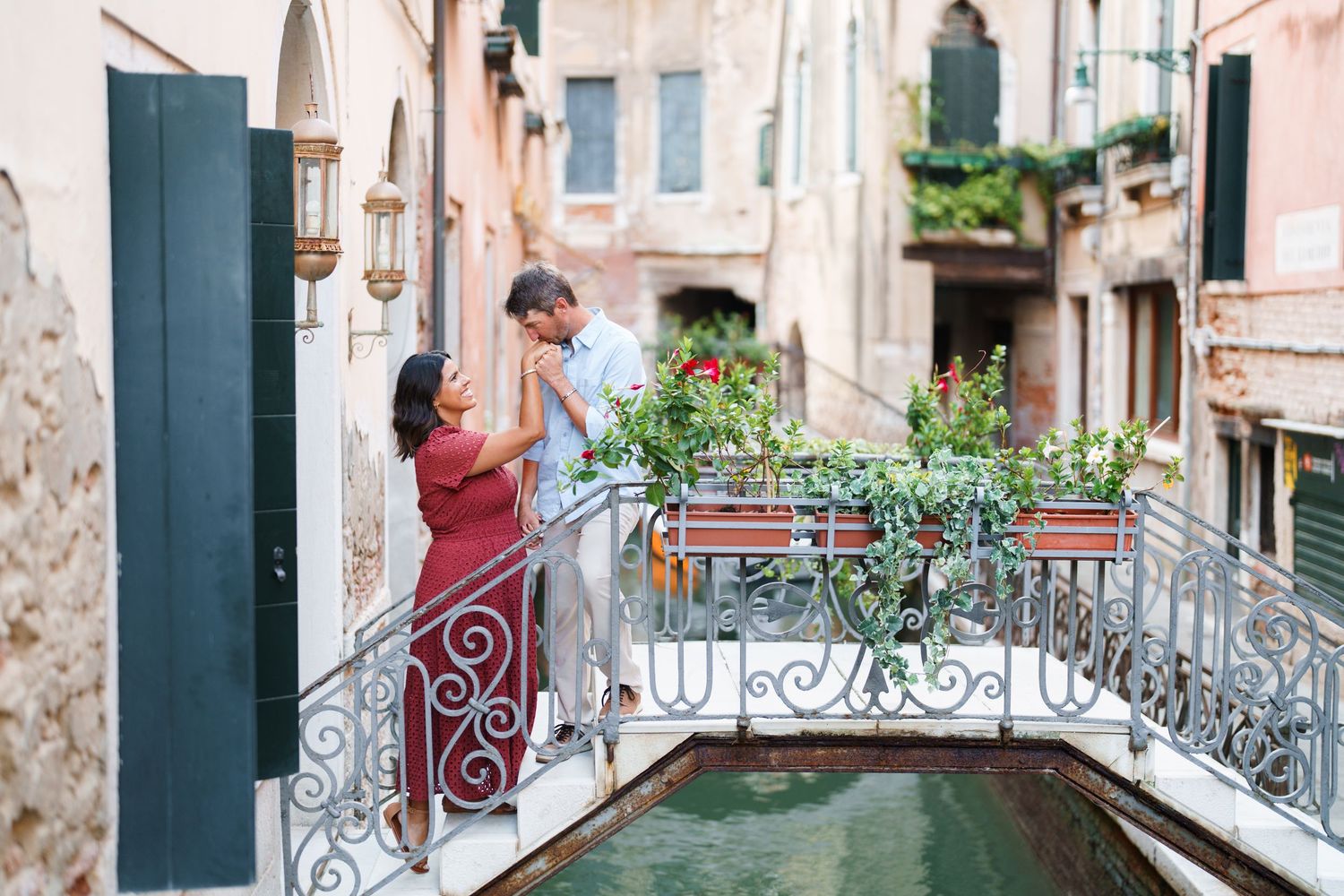 Couple sharing a romantic moment on a charming Venetian bridge over a canal, surrounded by colorful flowers.
