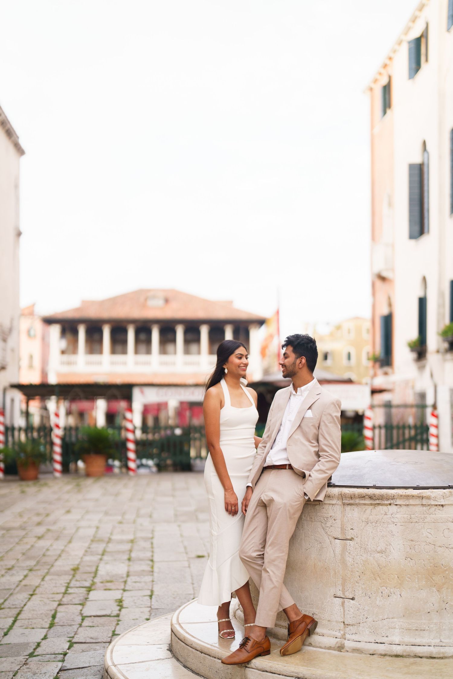 A couple in coordinated beige outfits sits together on stone steps in a European-style courtyard.