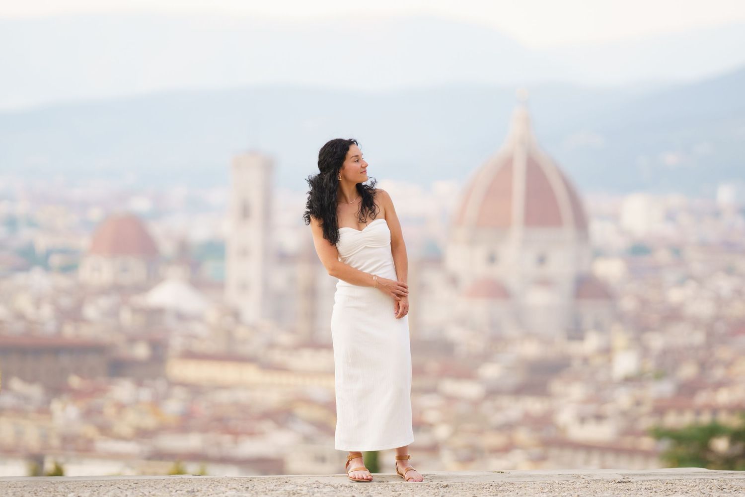 A person in a white dress poses against the scenic backdrop of Florence, Italy with the Duomo cathedral visible.