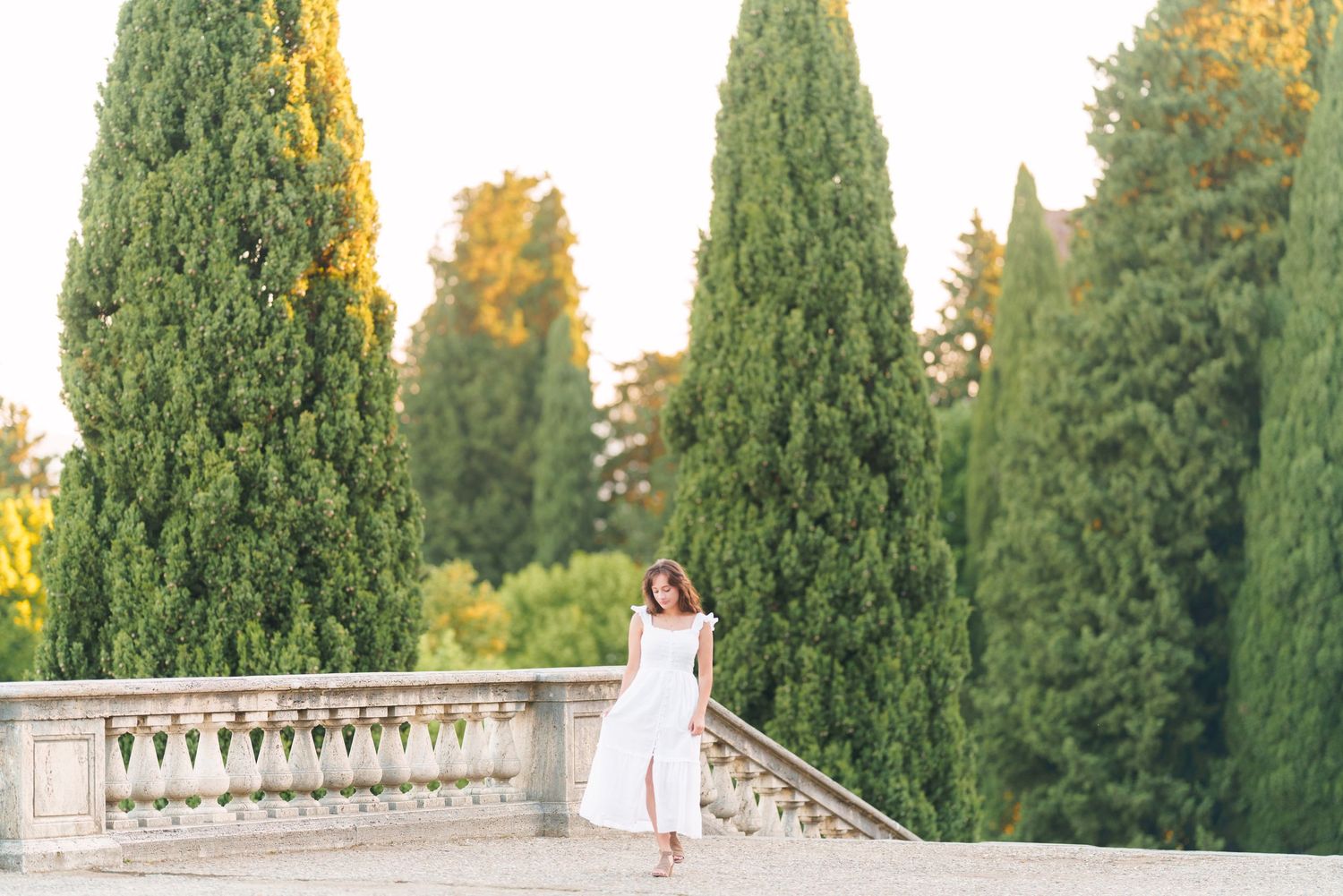 A bride in a white wedding dress stands on a stone balcony surrounded by tall cypress trees at sunset.