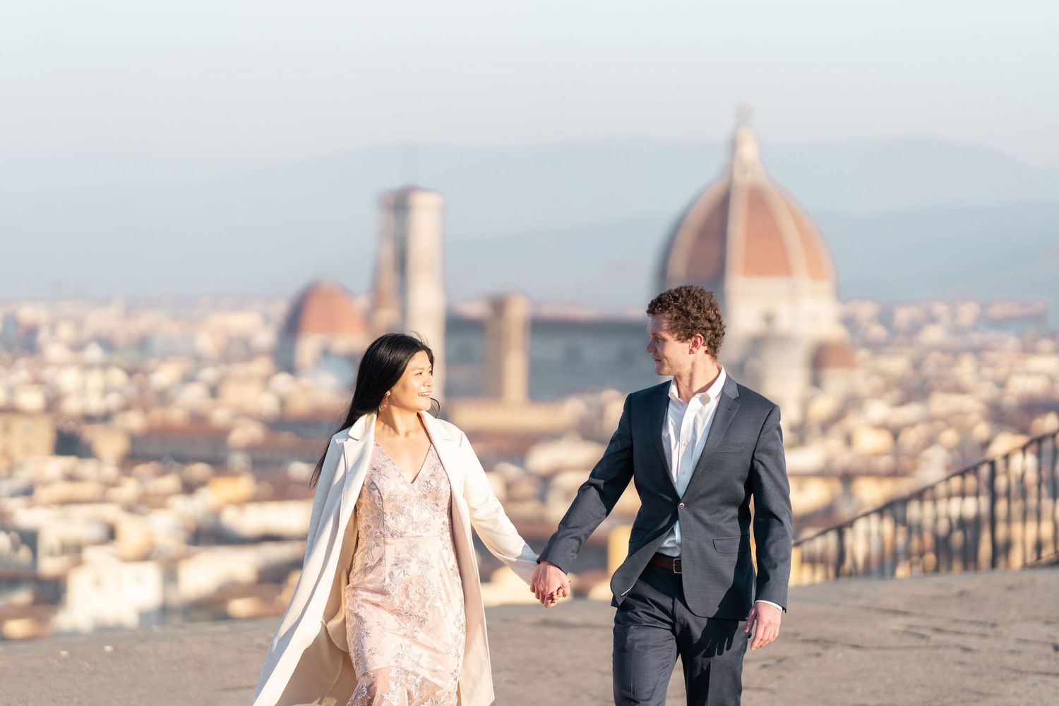 A couple holds hands while walking along a scenic overlook with Florence's Duomo cathedral visible in the background.