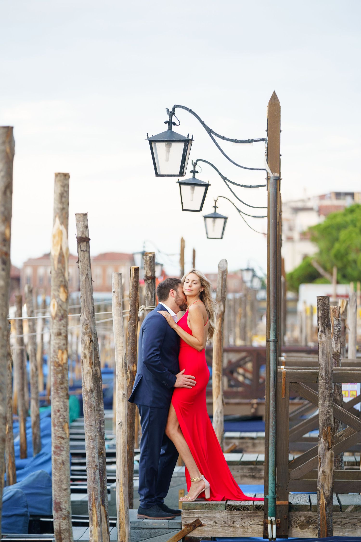 A romantic couple embraces near gondolas in Venice, with the woman wearing a flowing red dress against a waterfront backdrop.