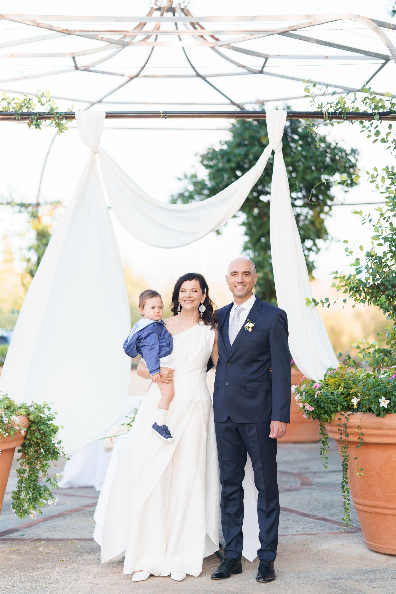 Wedding party poses under elegant white canopy with large terracotta planters in background.