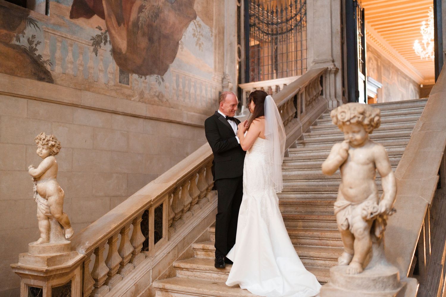 A romantic wedding moment on an elegant marble staircase adorned with cherub statues at a historic venue.