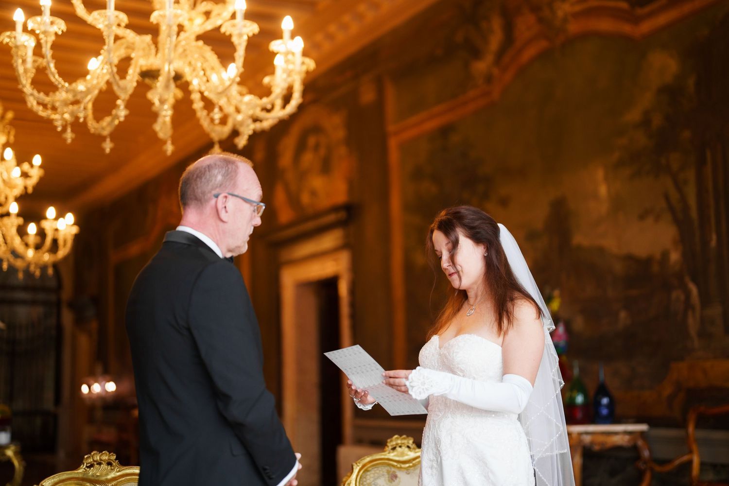 A couple exchanges vows during an elegant wedding ceremony in an ornate ballroom with crystal chandeliers.