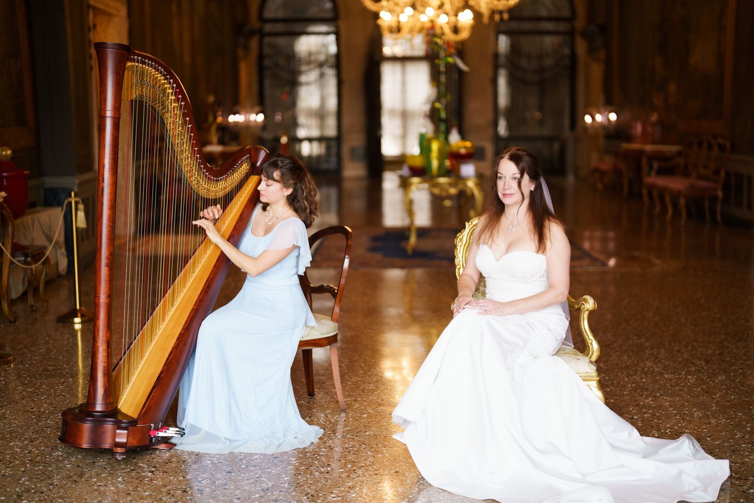 Two musicians in elegant white gowns perform with a classical harp in an ornate ballroom with chandeliers.