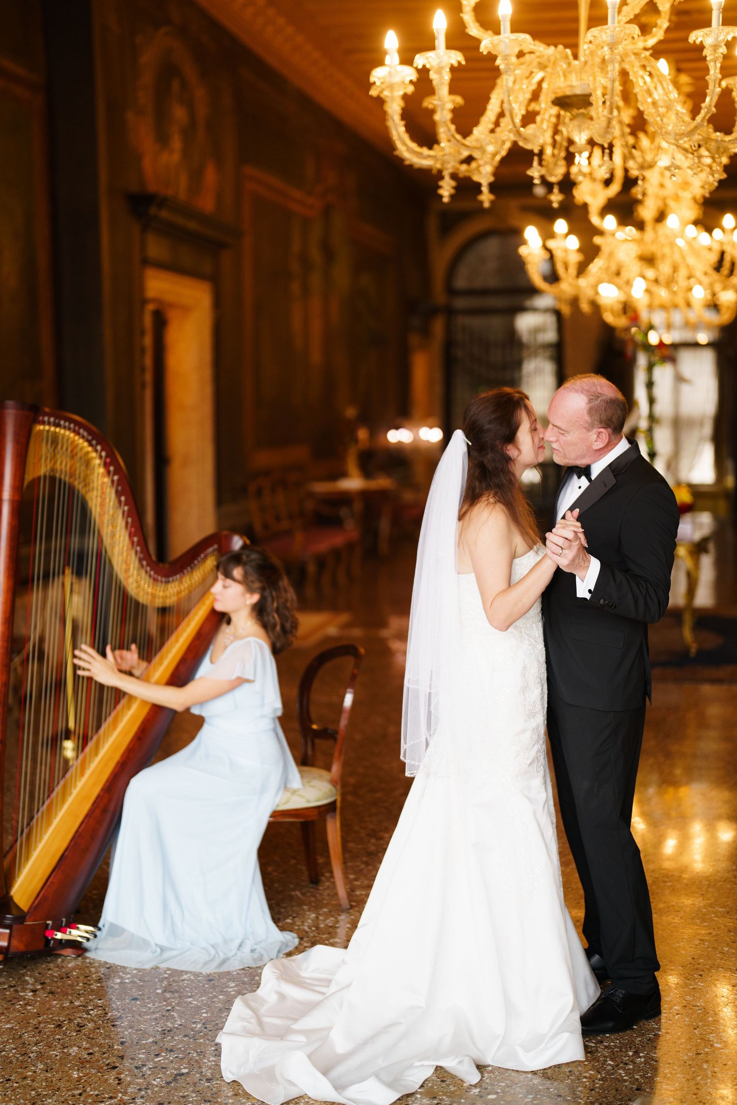Romantic wedding couple shares intimate moments in an elegant ballroom with crystal chandeliers and ornate harps.