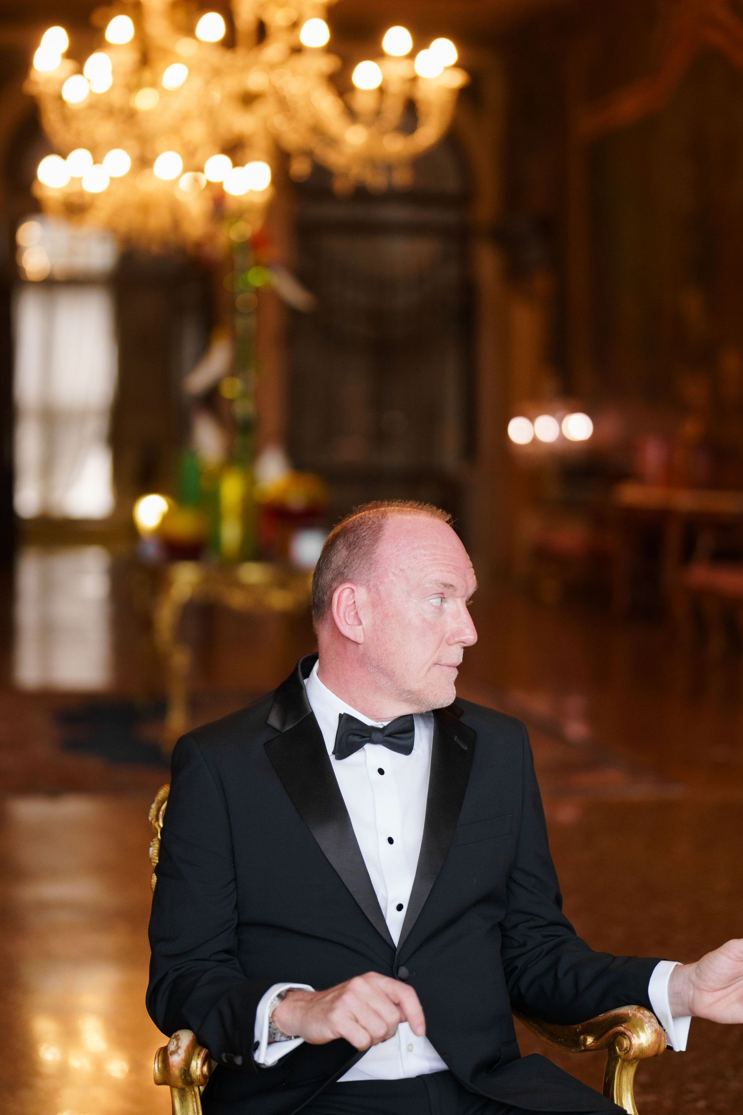 A person in a black tuxedo sits at a formal event with warm lighting and chandeliers in the background.