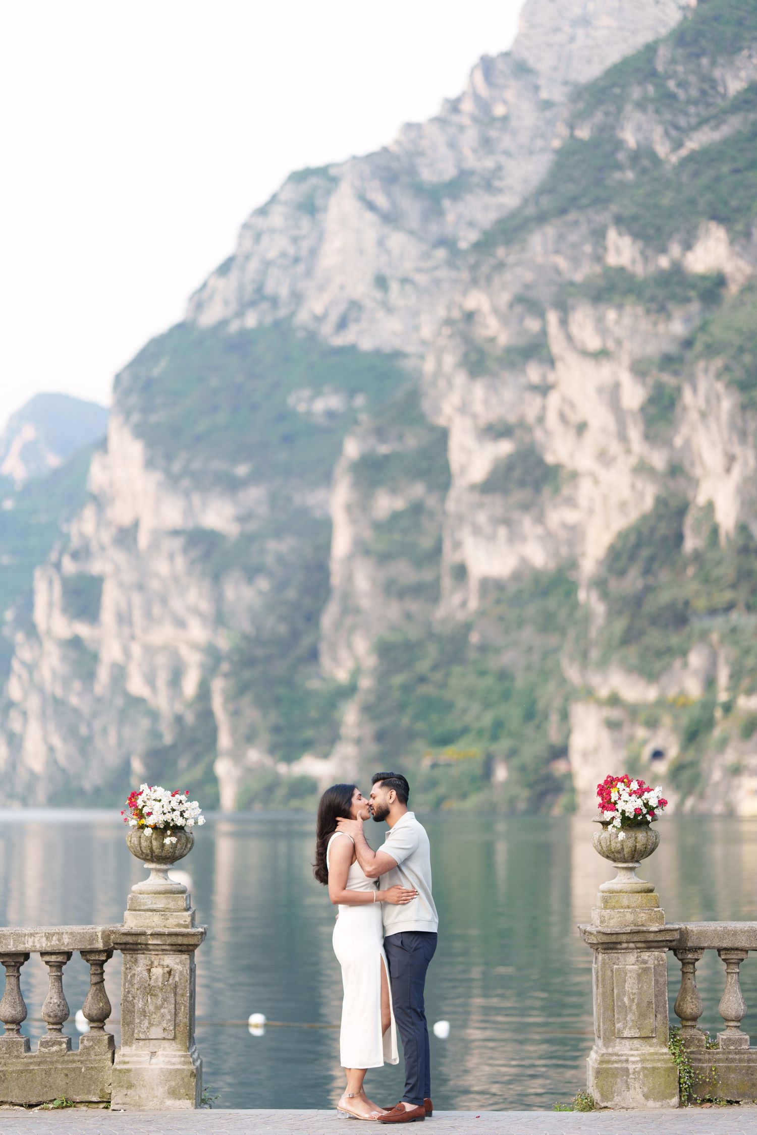 Romantic couple embraces on stone balcony overlooking scenic Lake Como with dramatic mountain cliffs in Italy.