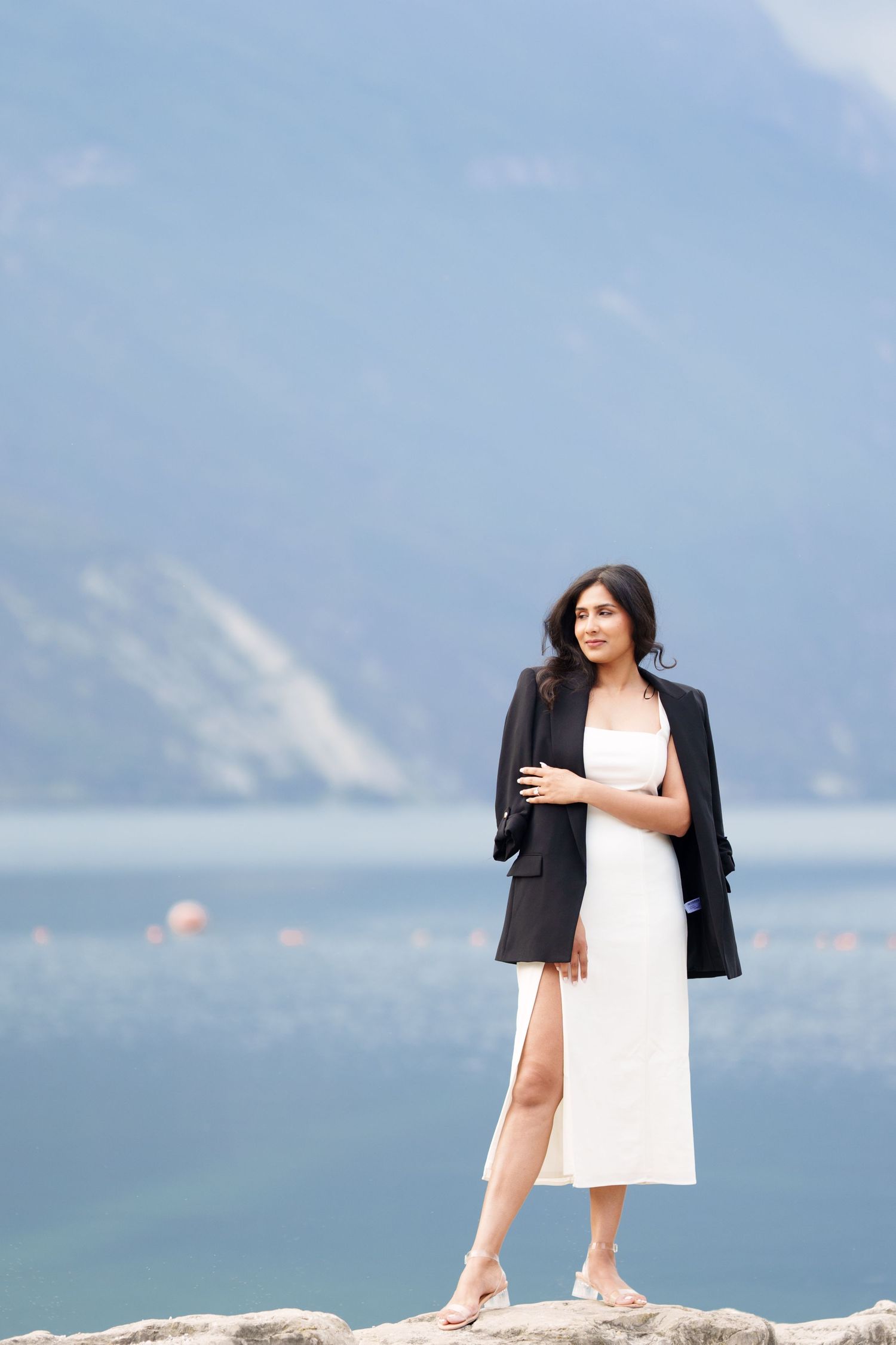 A person in a black blazer and white dress stands on a rocky overlook with mountains and water in the background.