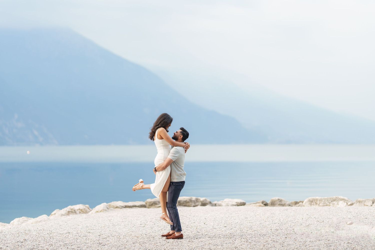 Two people embrace on a scenic beach with mountains and water in the background during a romantic moment.