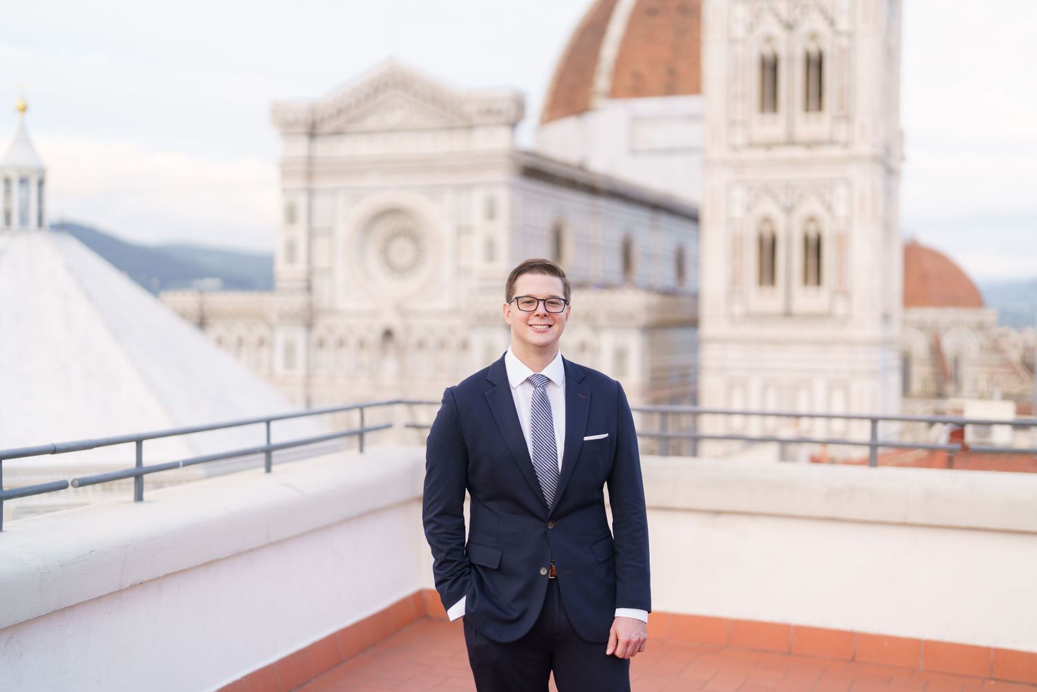A person in a navy suit stands on a rooftop terrace with ornate stone architecture and domed buildings in the background.