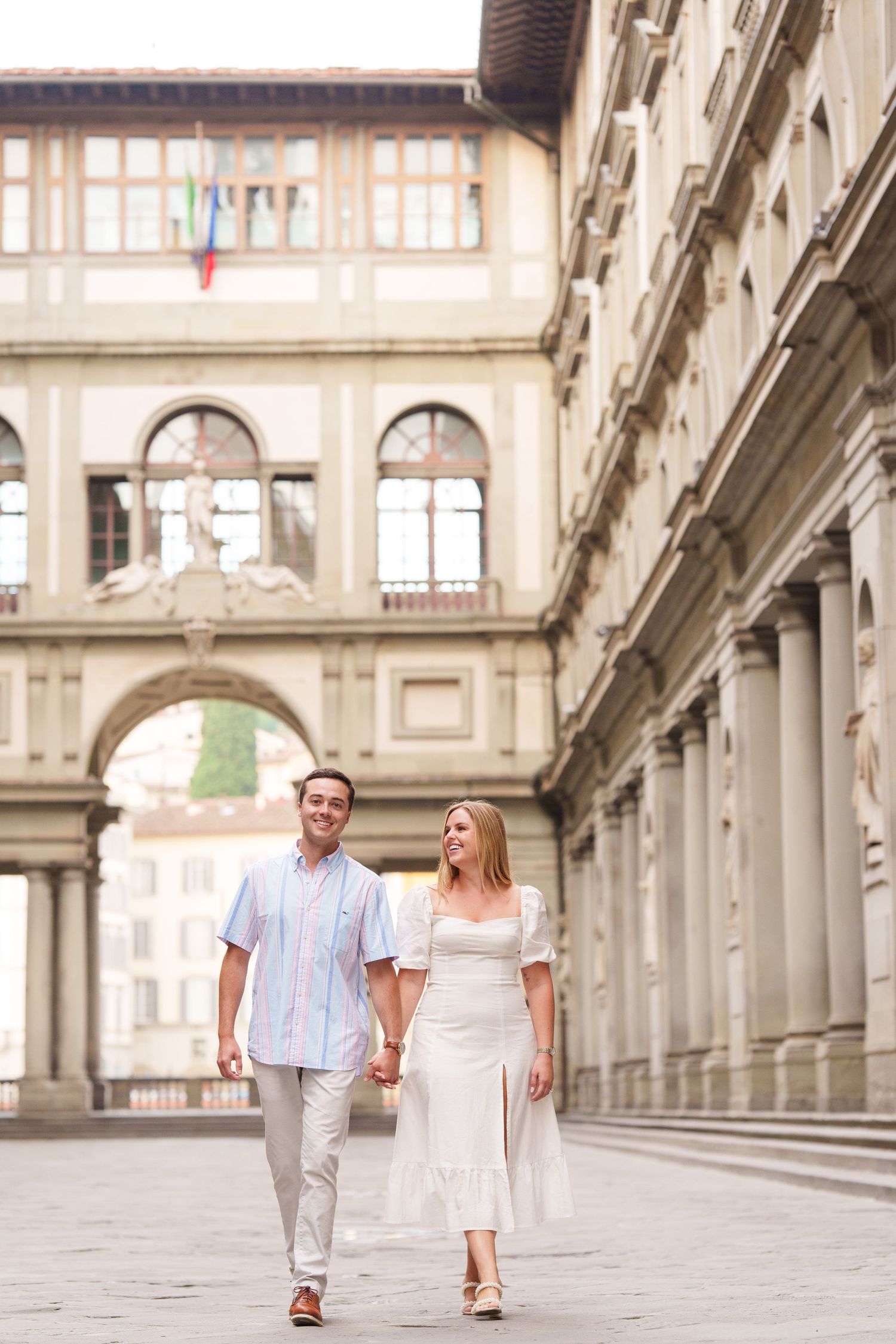 A couple walks hand in hand through an elegant European colonnade with ornate architecture and natural lighting.