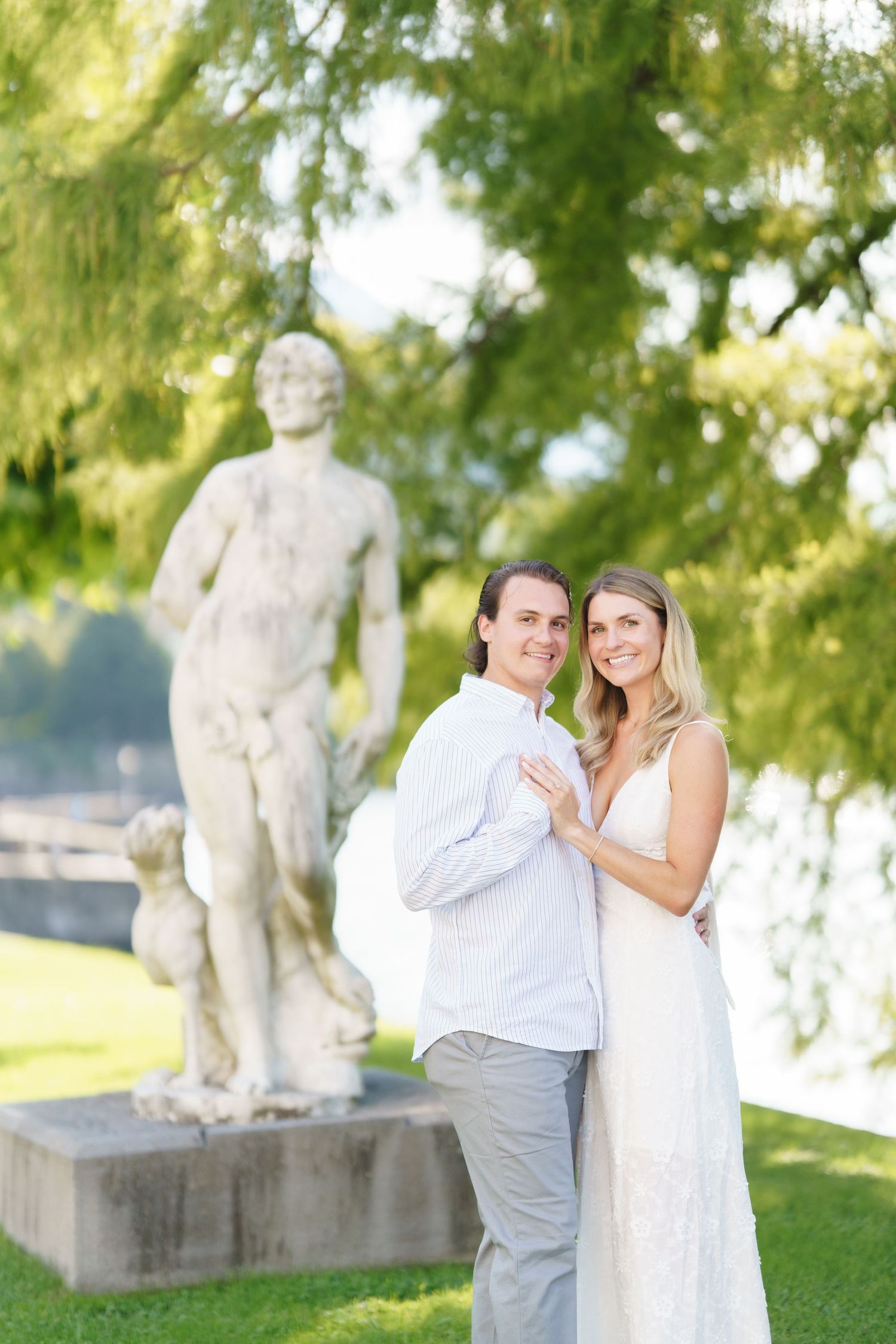 A couple poses together on a sunny day near a classical stone statue in a garden setting.